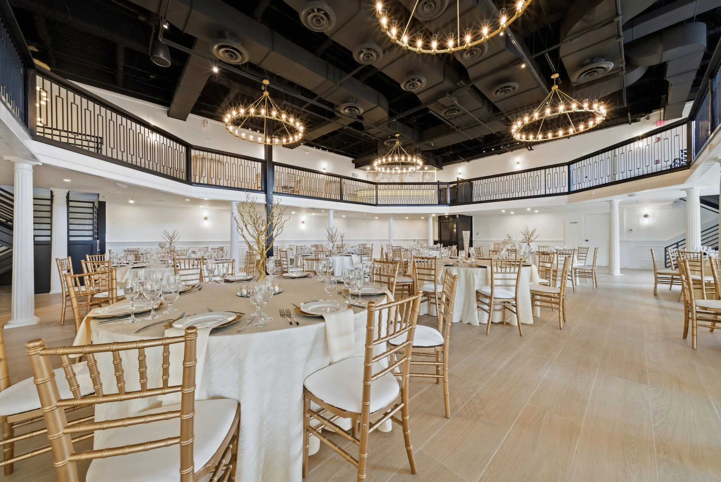 Elegant banquet hall with round tables covered in cream tablecloths, set with glassware, silverware, and white plates, surrounded by gold Chiavari chairs. The hall features a high ceiling with black exposed ducts, drum chandeliers with numerous small