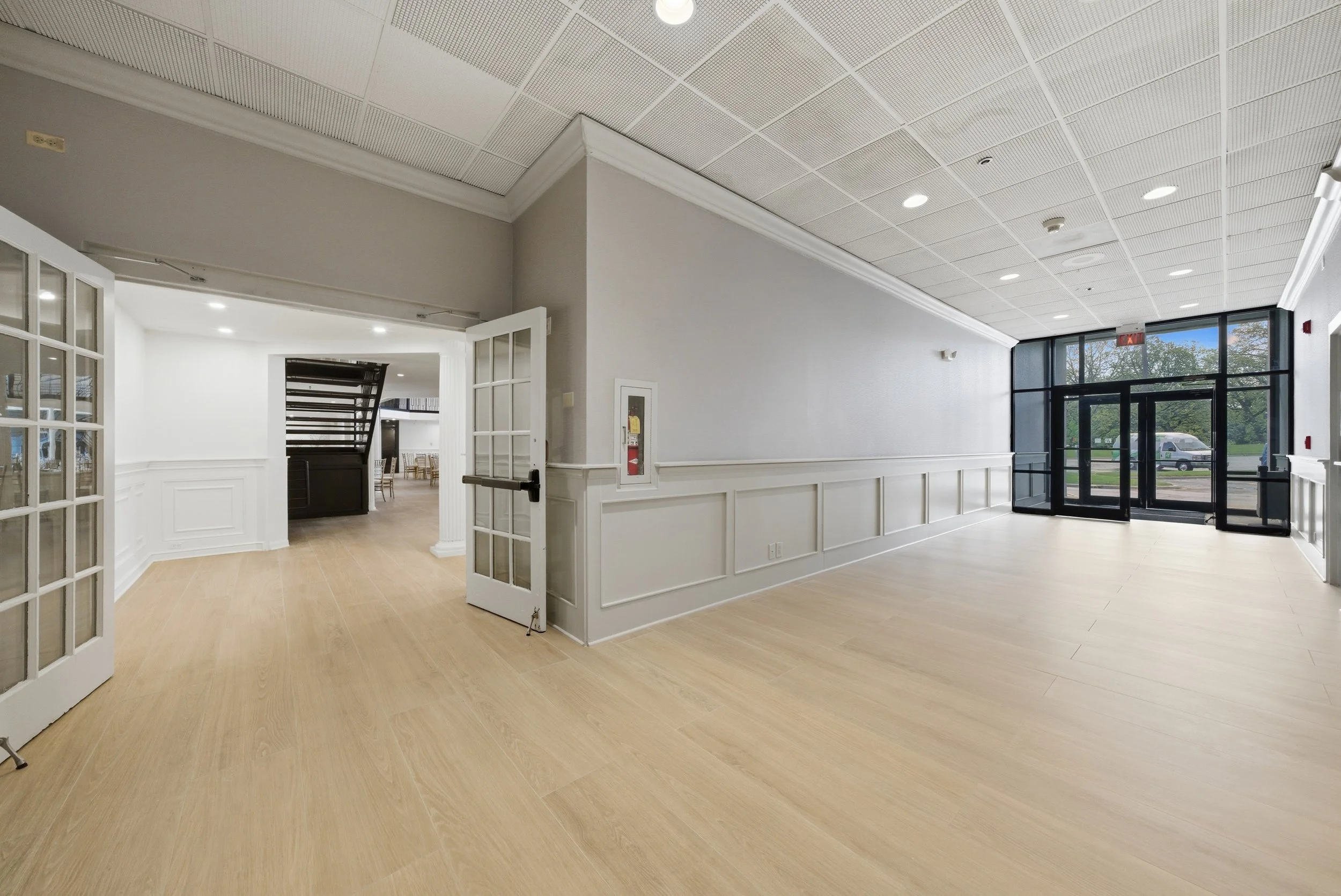 Interior of a modern building with beige wood flooring, white walls, a drop ceiling with recessed lighting, and large glass doors leading outside. Some furniture is visible through an open doorway.
