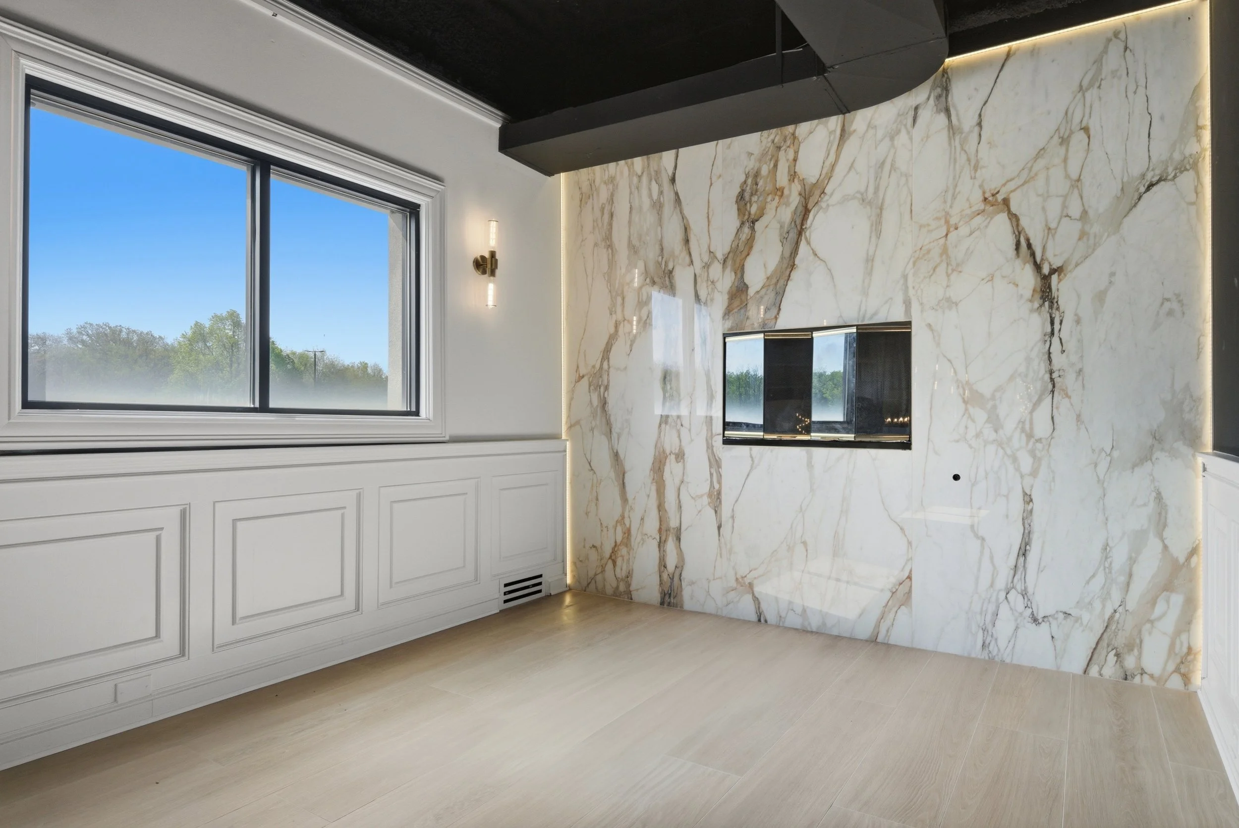 Interior of a modern room featuring a large window with a scenic view of trees and a blue sky, white wainscoting on the lower wall, a marble accent wall with gold and black veining, and a fireplace inset in the marble wall. The room has a minimalist 