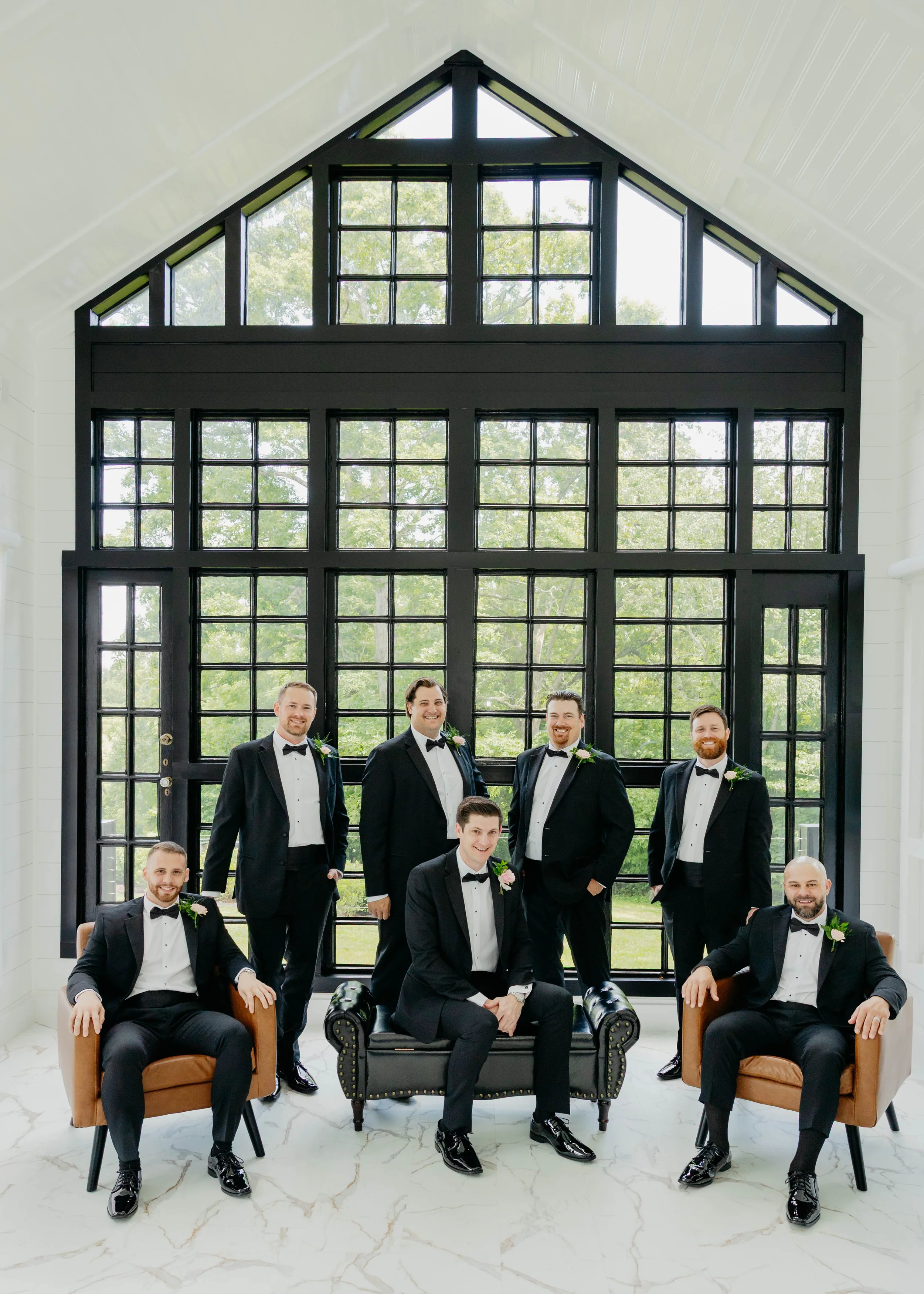 A group of nine men in black tuxedos with white shirts and black bow ties, posing in front of large black-framed windows with greenery outside. They are smiling and some are sitting while others are standing, indoors with a white marble floor and whi