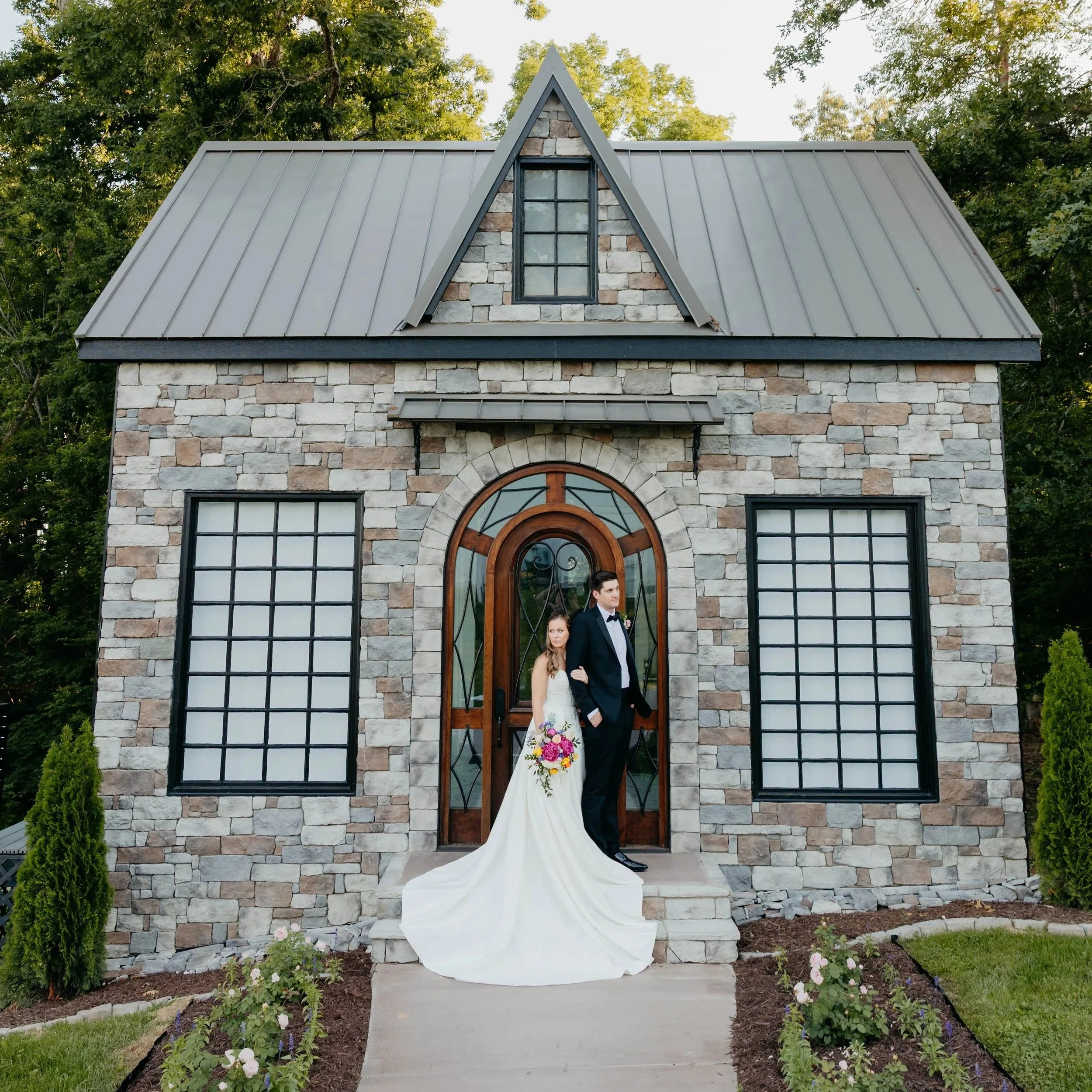 A wedding couple standing at the entrance of a stone English Garden Glass Cottage with large windows, a wooden door, and a steep gabled roof, surrounded by green trees and colorful flowers.
