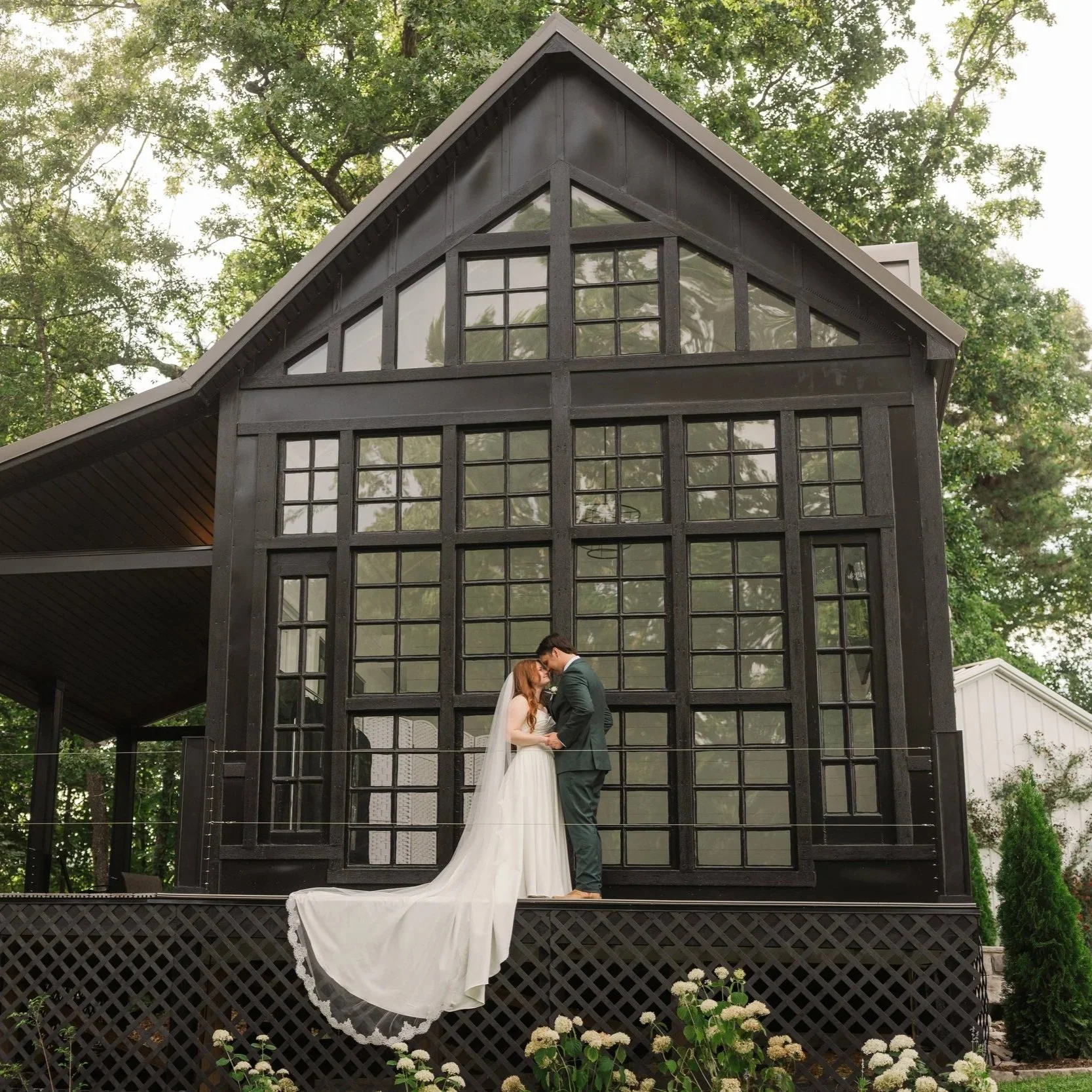 A bride and groom standing close together, holding hands, and tilting their foreheads together in front of a modern black glass cottage with multiple windows, surrounded by trees and flowers.