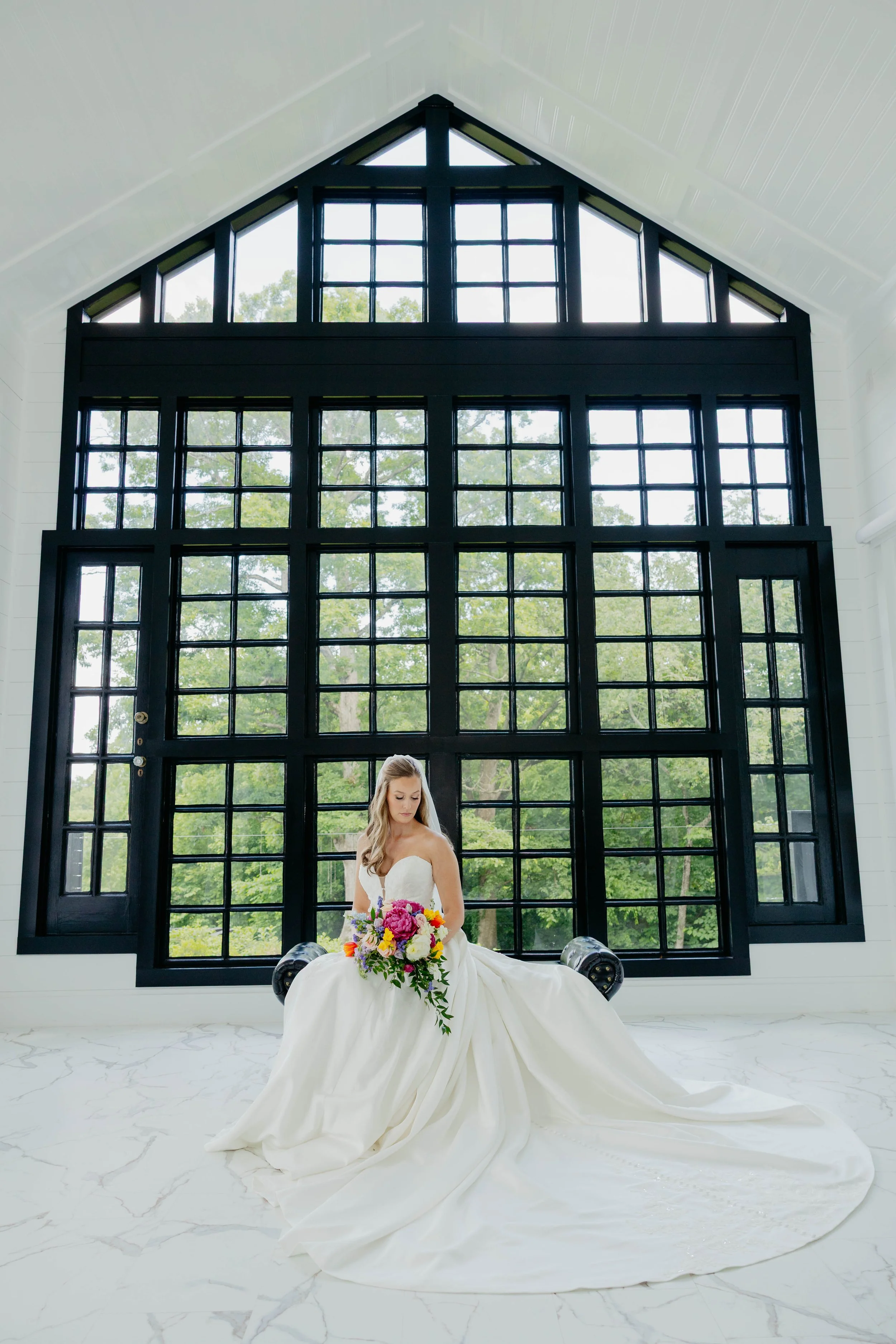 A bride in a white wedding gown sitting on a black bench, holding a bouquet of colorful flowers in front of a large black geometric window, with a white marble floor and green trees visible outside.