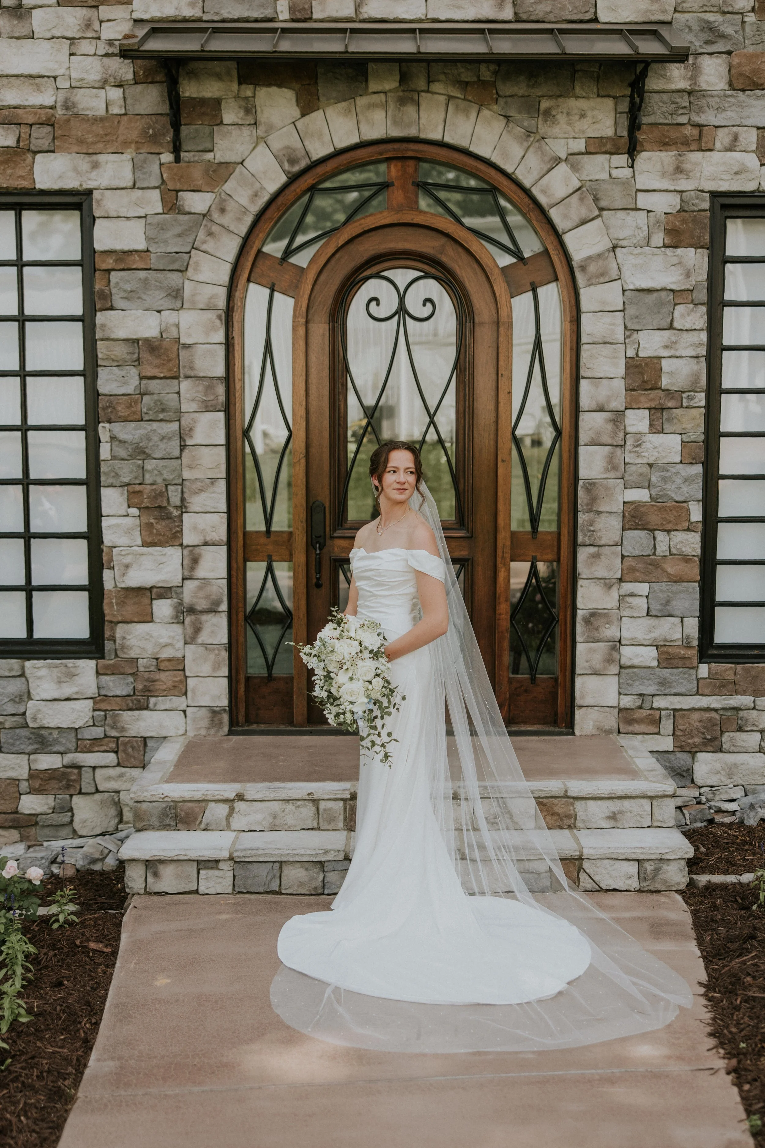 A bride in a white wedding dress holding a bouquet of flowers standing on the steps in front of an English Garden Glass Cottage with stone front and elegant round wooden door.