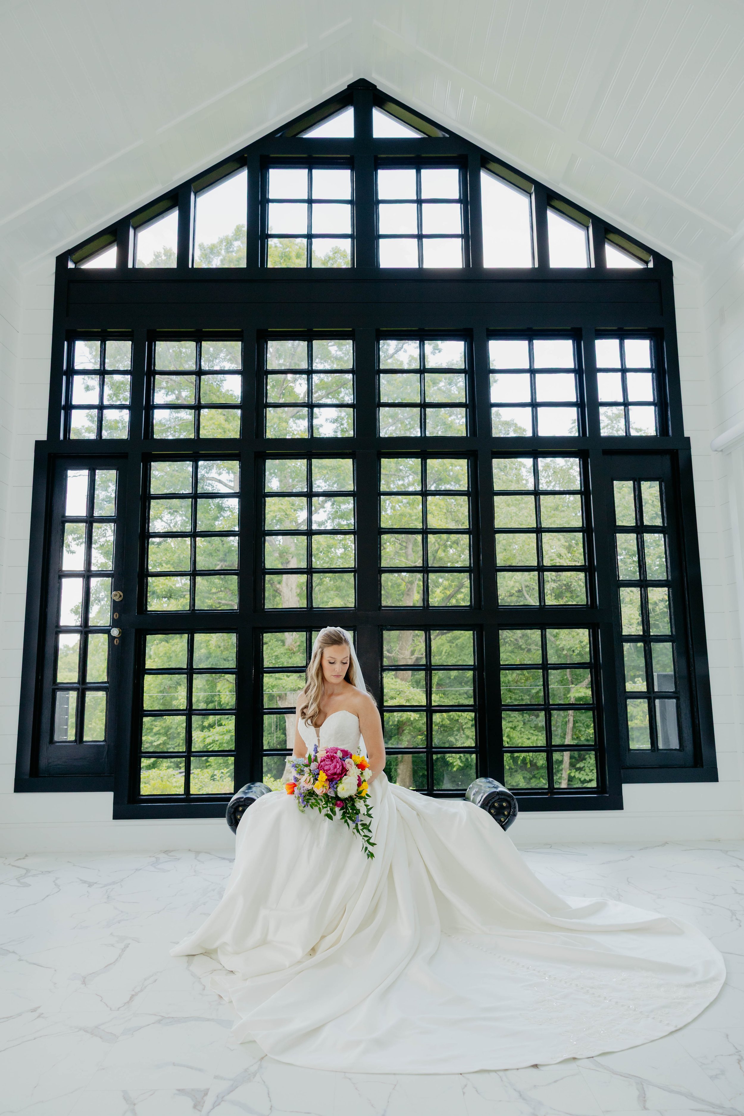 A bride in a strapless white wedding gown sitting on a bench in front of a large black-framed window with multiple panes, holding a colorful bouquet of flowers, with a scenic view of green trees outside.