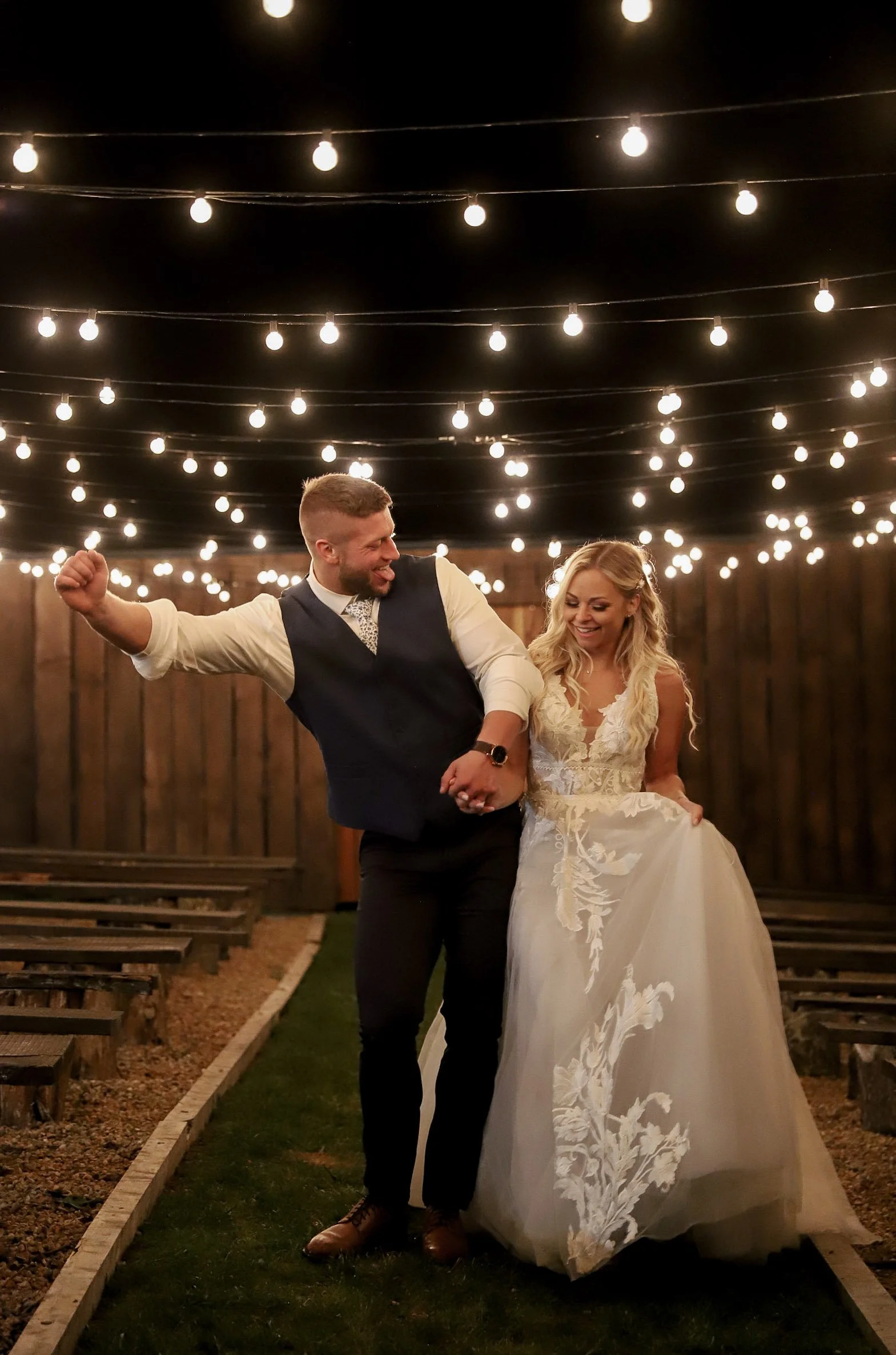 A bride and groom stand under a wooden wedding arch decorated with pink and white flowers and greenery, outdoors with mountains and trees in the background.
