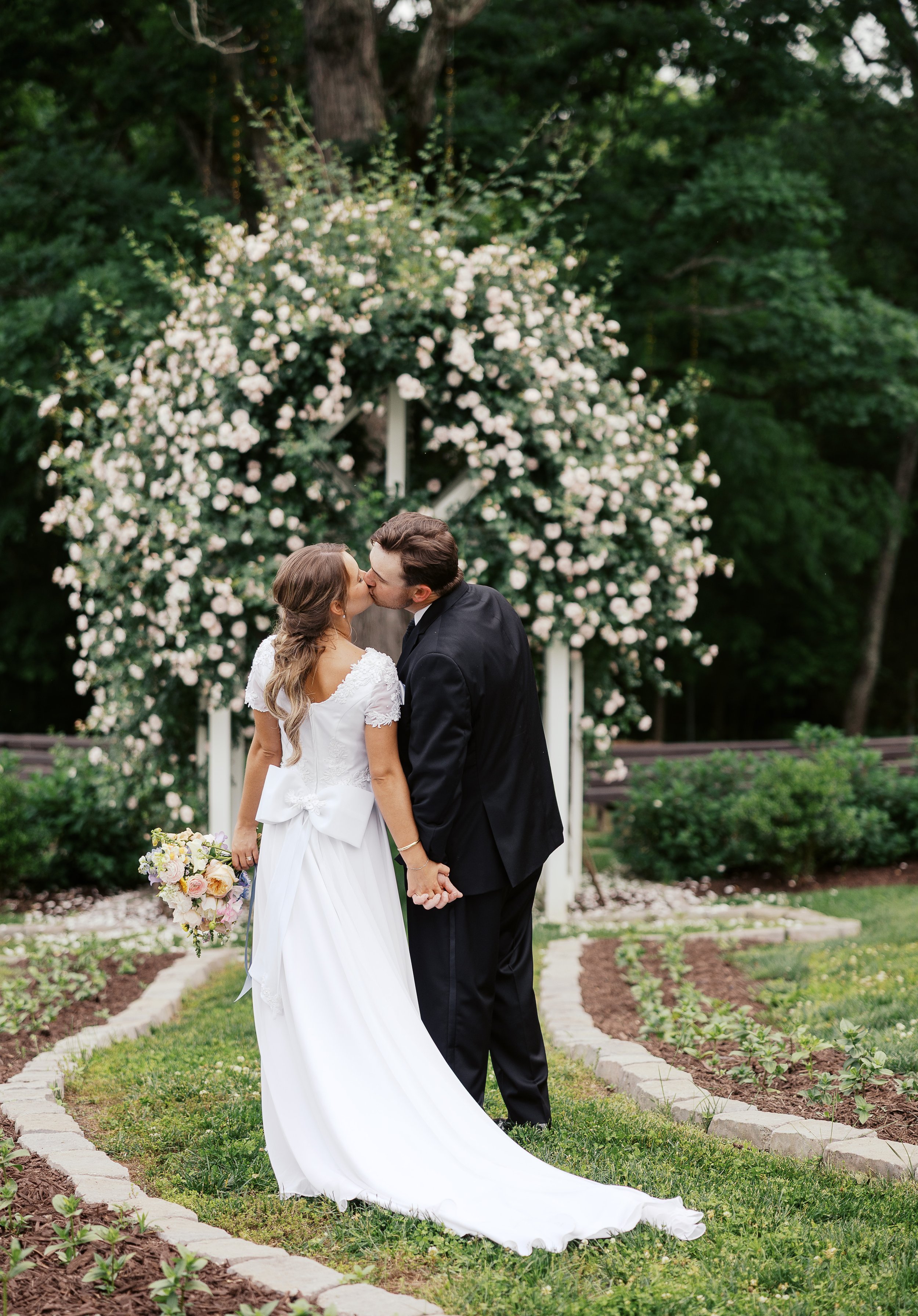 A newlywed couple sharing a kiss outdoors, holding hands, with a floral arch and greenery in the background.