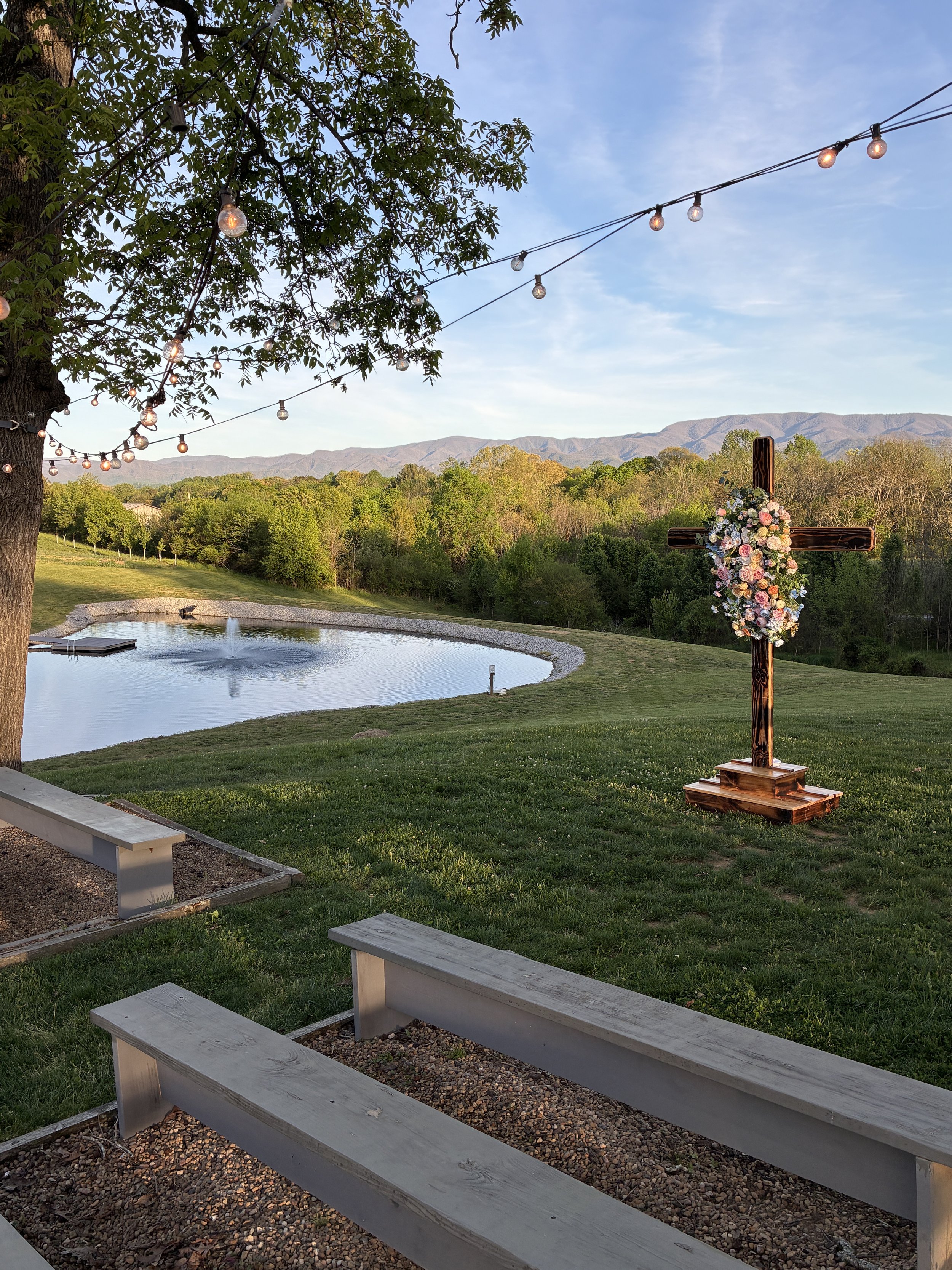 Outdoor ceremony site with string lights hanging from a large tree, a wooden cross decorated with flowers, a pond with a fountain, and a the smoky mountains in the background during early evening.