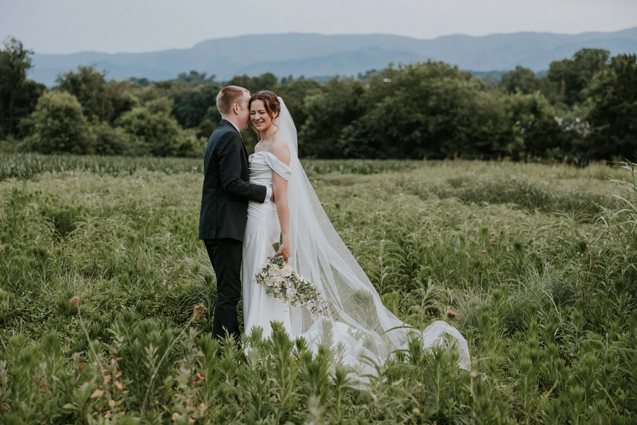 A bride and groom standing together in a green field, smiling and embracing each other, with mountains and trees in the background.