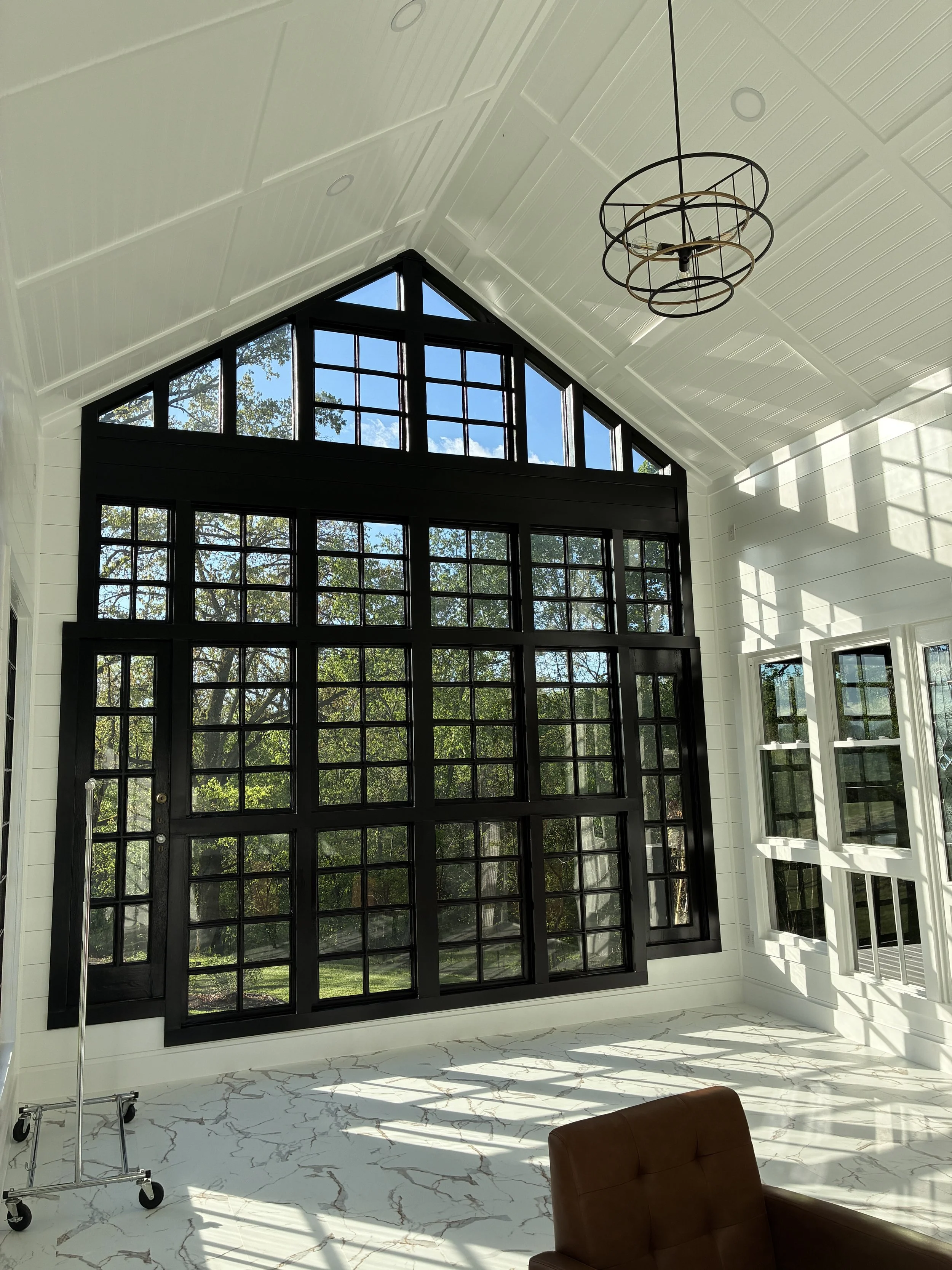 Interior of a bright room with large black-framed windows, white shiplap walls, a white ceiling, and a modern hanging chandelier. Sunlight streams through the windows, casting shadows on the marble floor, with a brown leather chair visible in the for