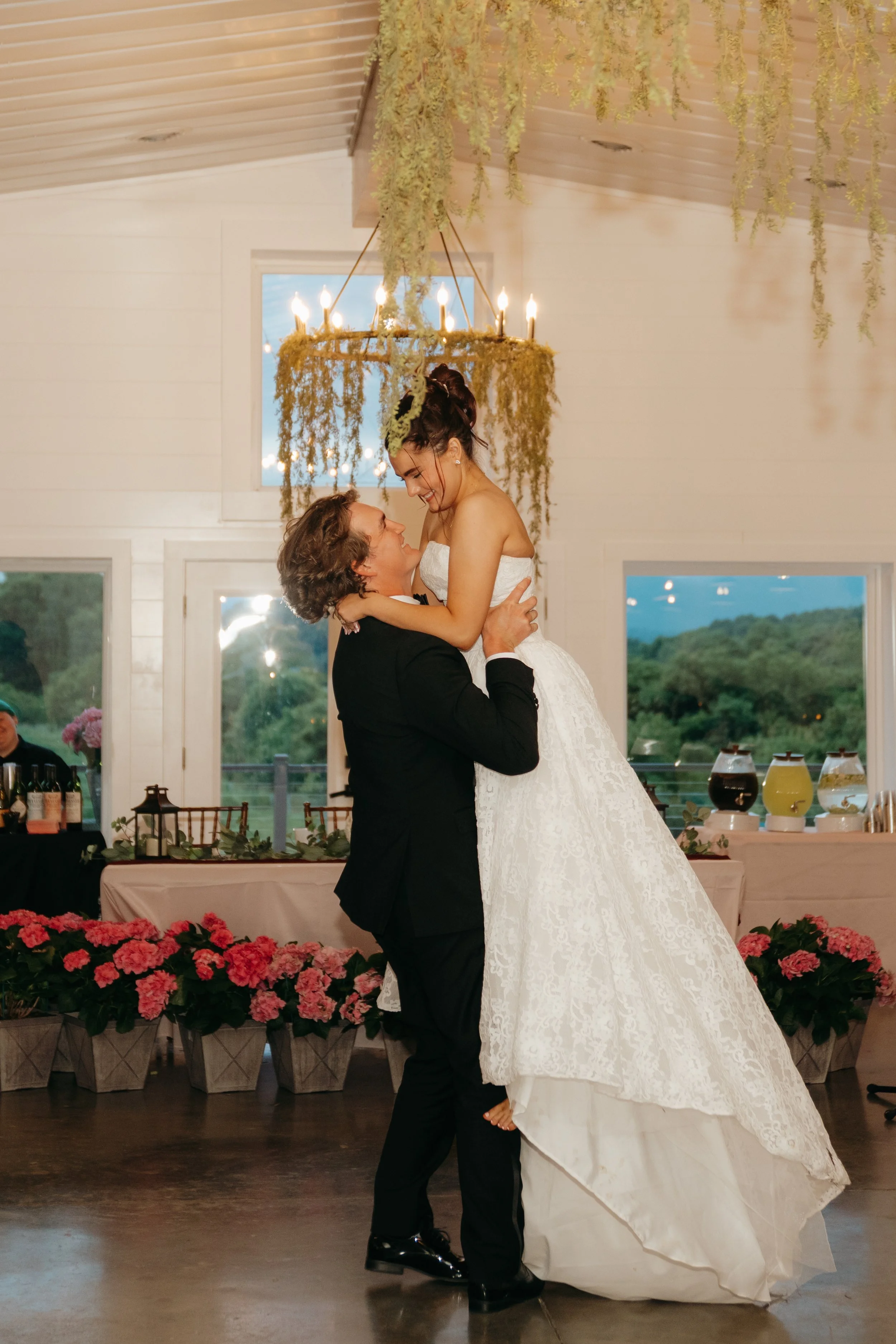 A bride and groom dancing at their wedding reception inside a decorated venue with large windows, hanging greenery, and a floral arrangement of pink flowers along the floor.