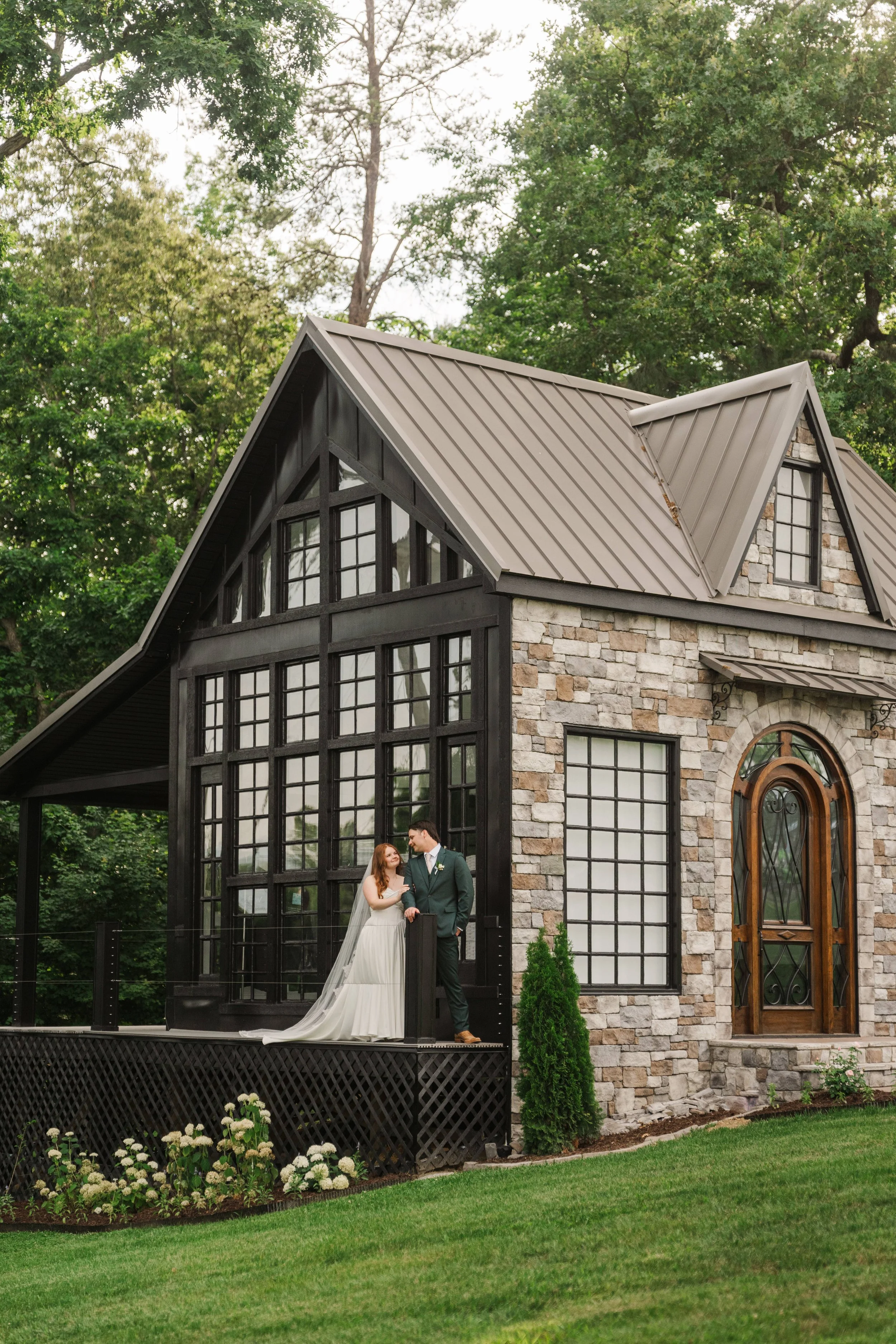 A newlywed couple stands on a deck in front of a modern house with large windows and stone walls, surrounded by greenery, trees, and flowers.