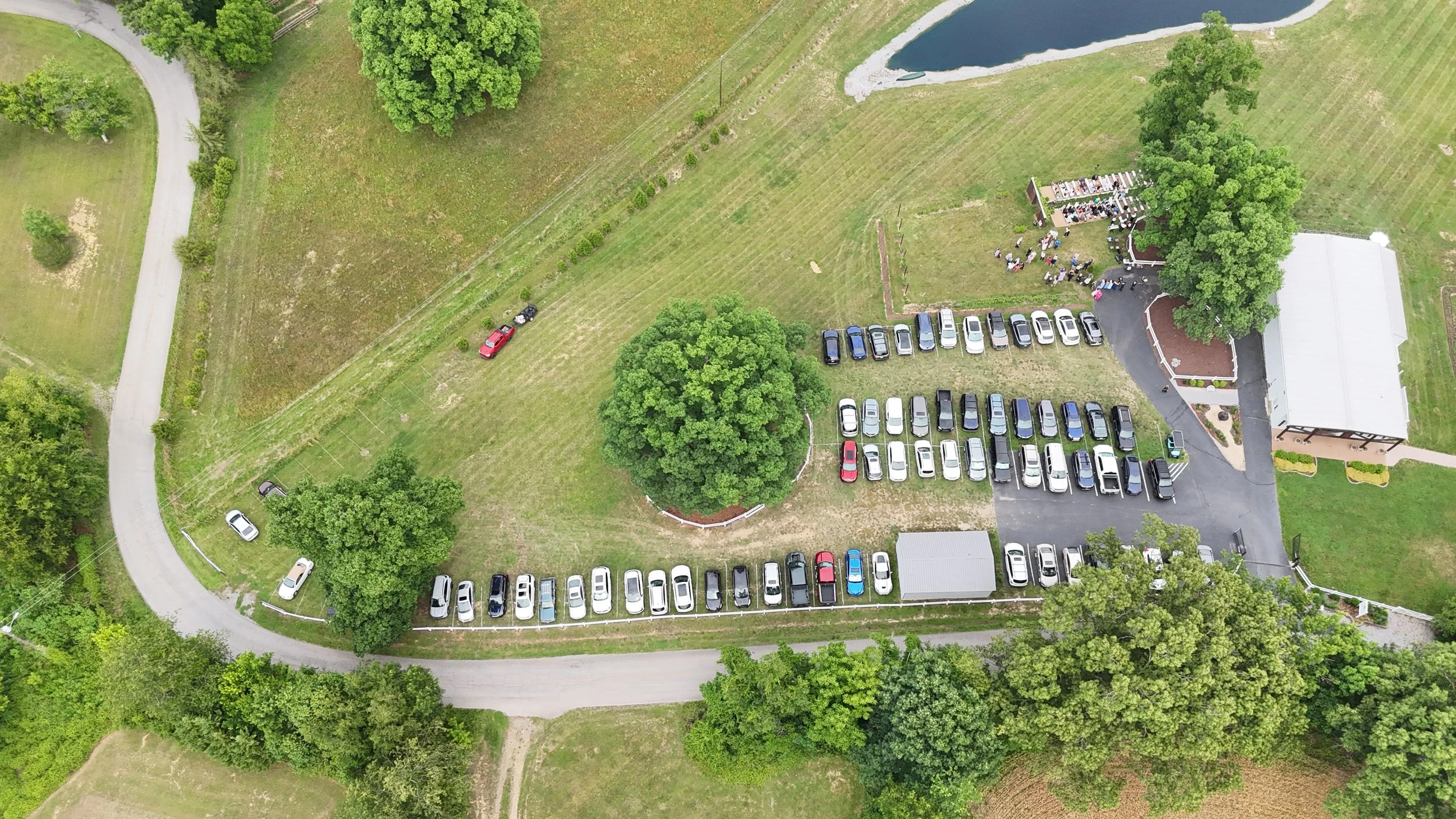 An aerial view of a rural area with a parking lot filled with cars, a white building, lush green trees, and a small pond. There are people gathered near the building.
