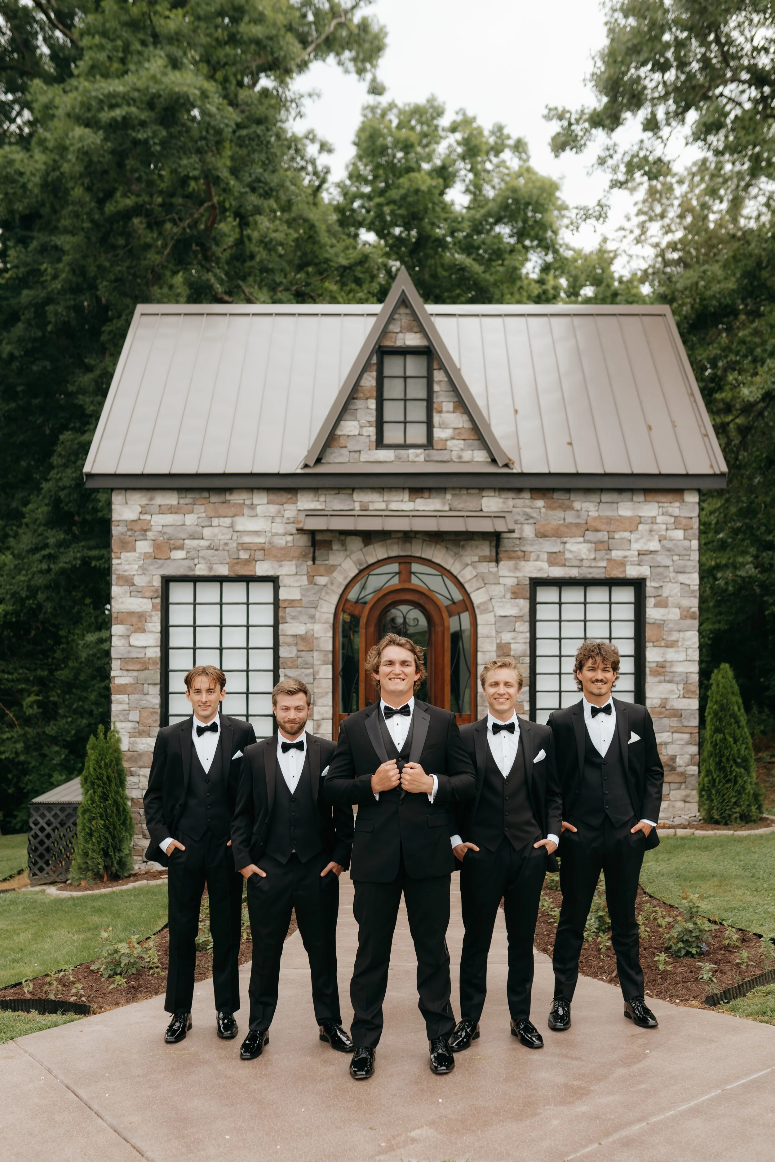 A group of six men in black tuxedos and bow ties standing outdoors in front of a stone glass cottage with a steep metal roof and large windows, smiling and posing for the camera.