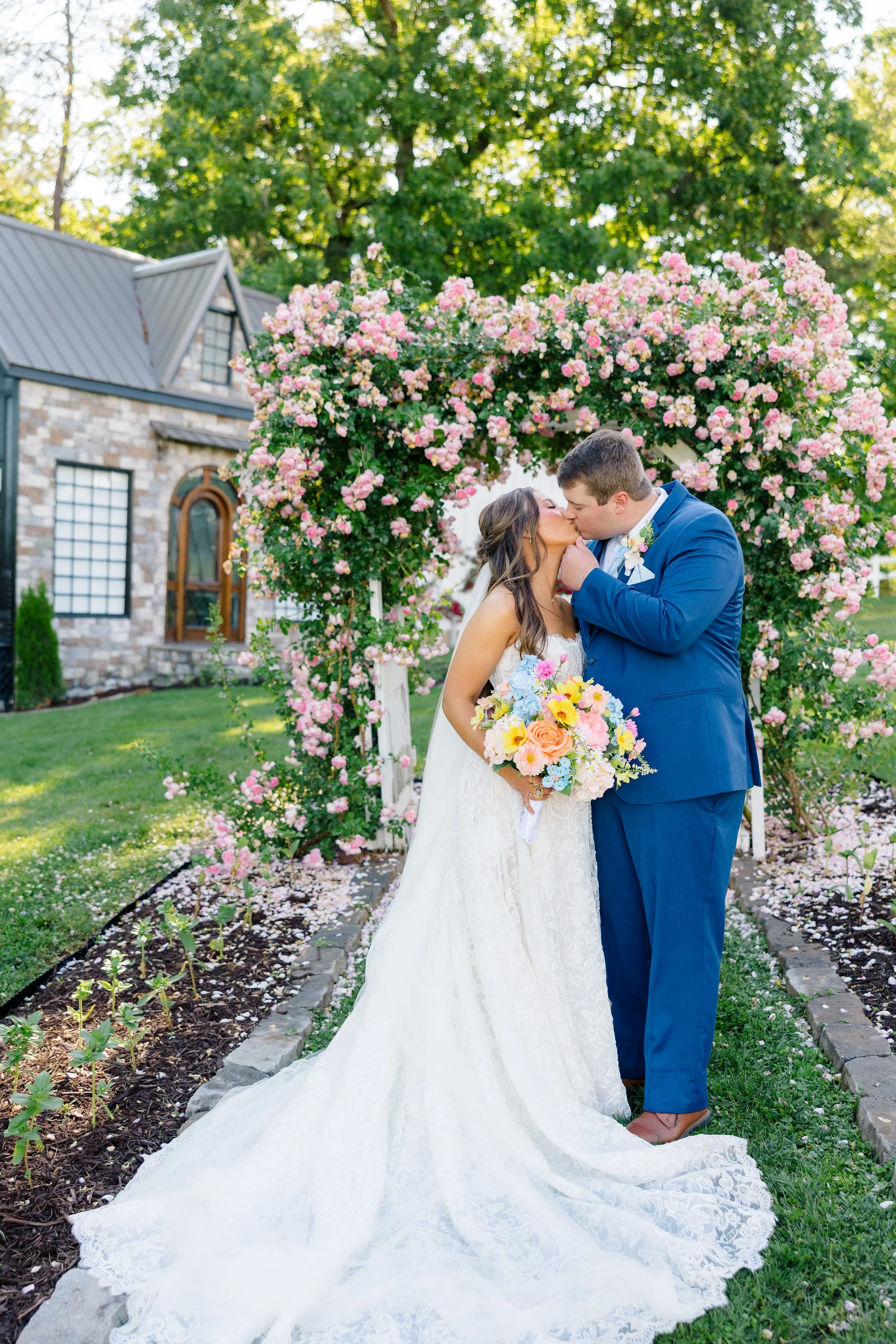 A bride and groom kissing under a pink flowering arch outdoors during a wedding ceremony.
