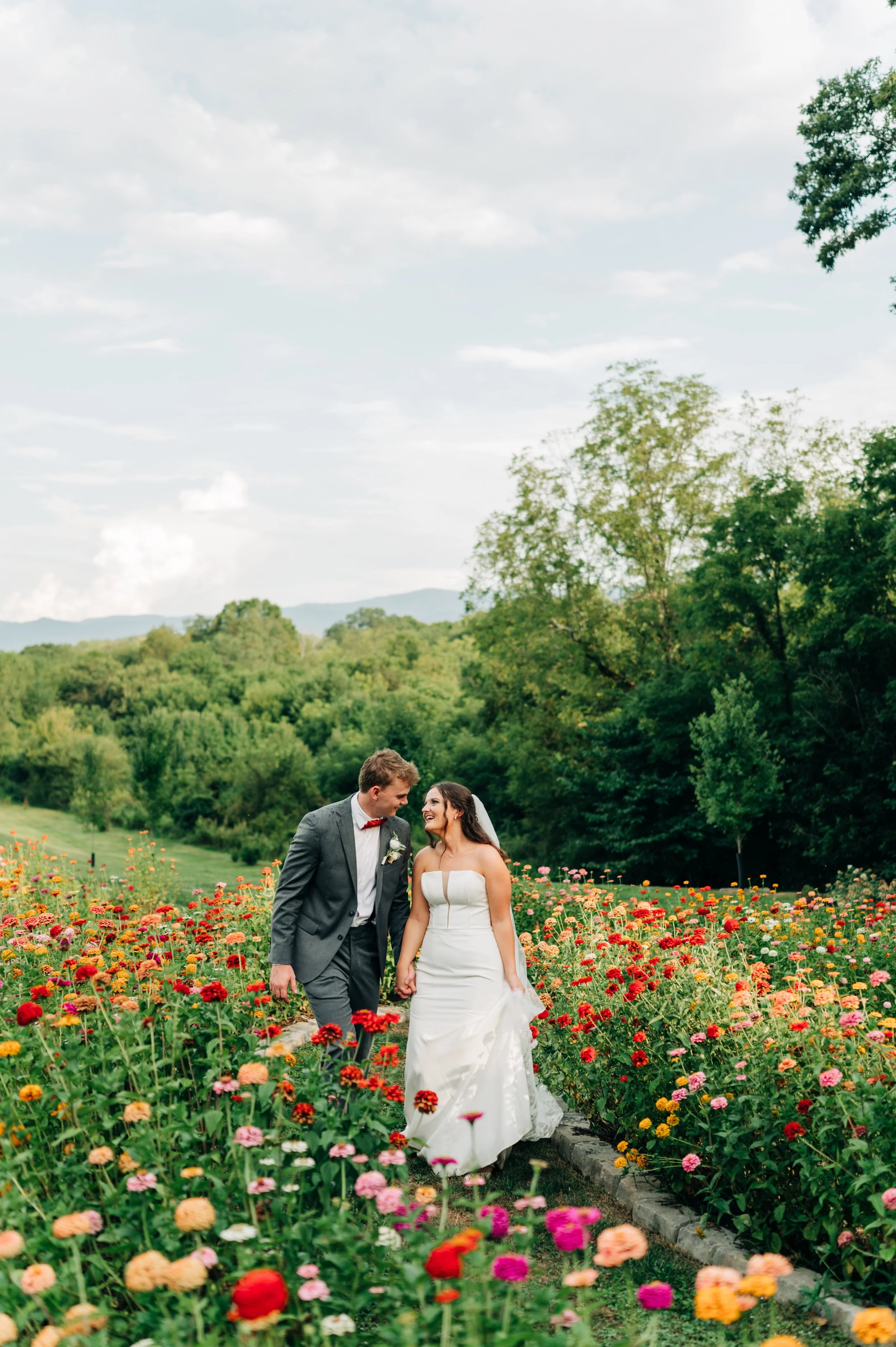 A bride and groom walking hand in hand through a colorful flower garden, smiling at each other.