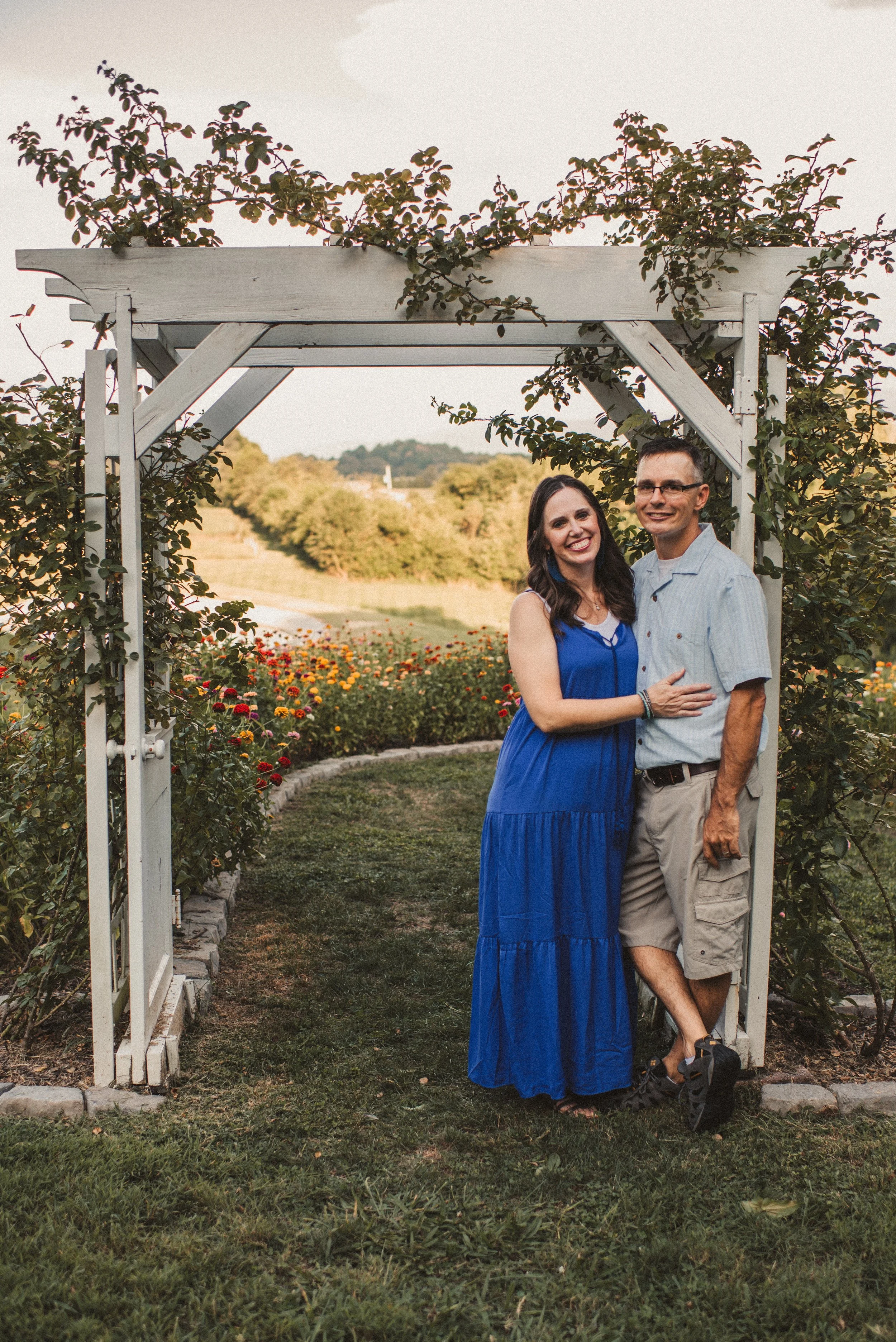 A smiling couple stands under a white garden arbor with climbing plants in a scenic outdoor setting, with a flower bed and rolling hills in the background during sunset.