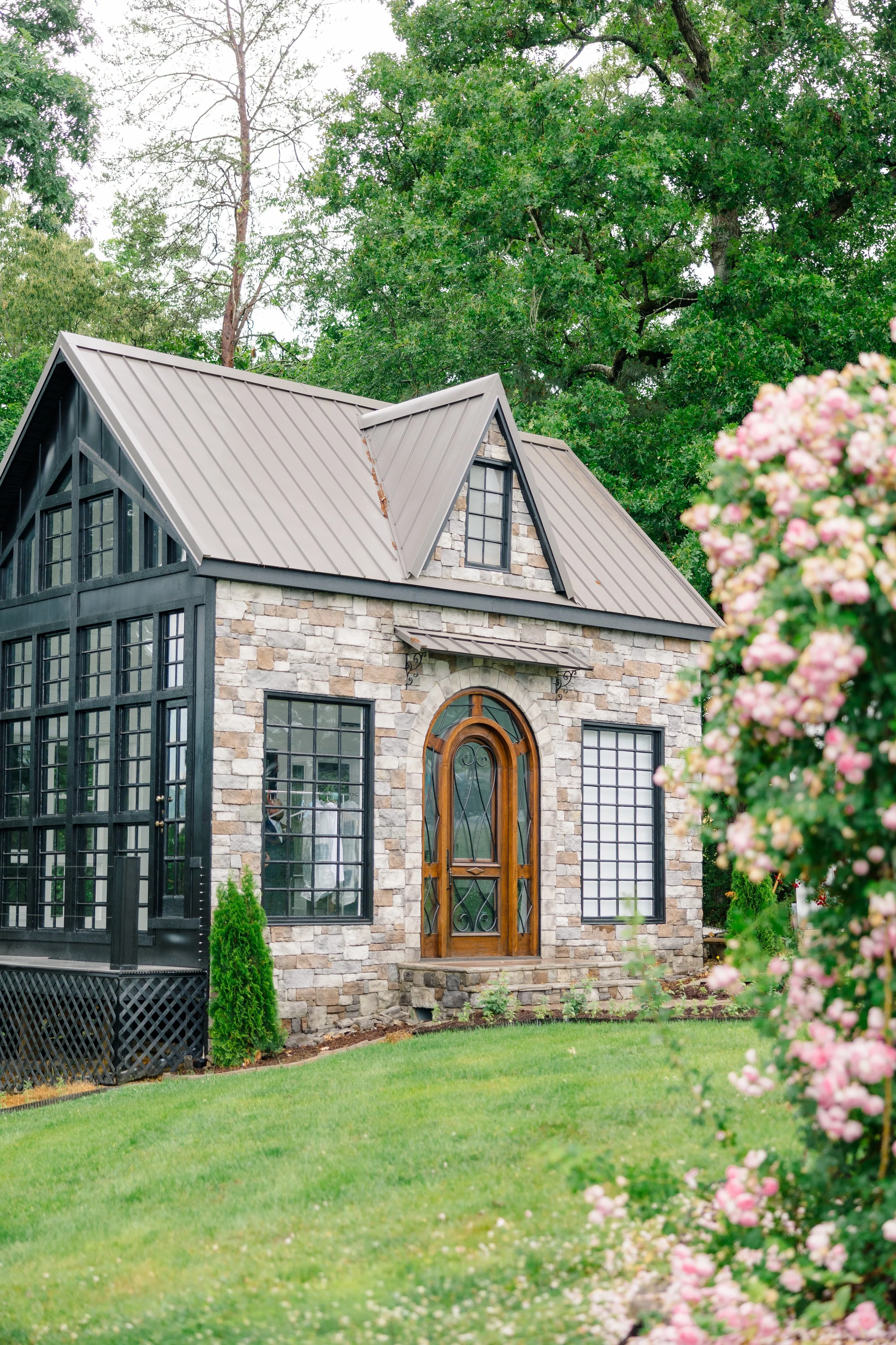 A glass cottage with a stone exterior, large windows, and a wooden arched door, surrounded by a well-maintained lawn and lush greenery, including large trees and flowering roses.