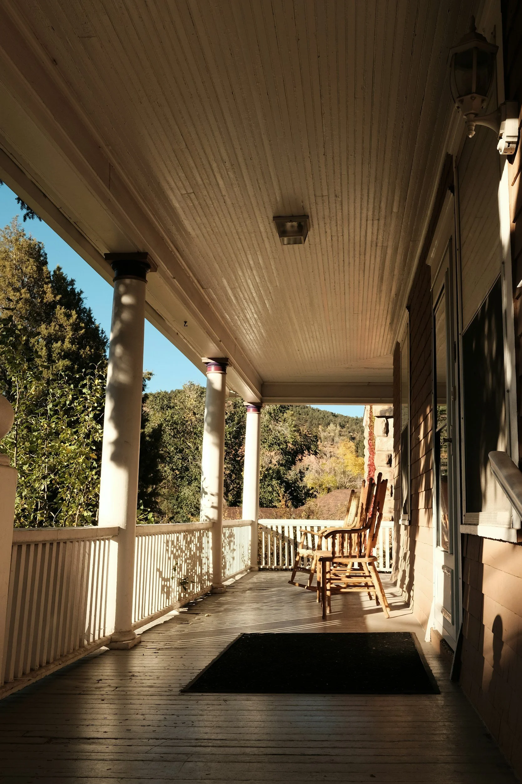 A porch with a visible doormat and two chairs. The sun is shining and casting long shadows.