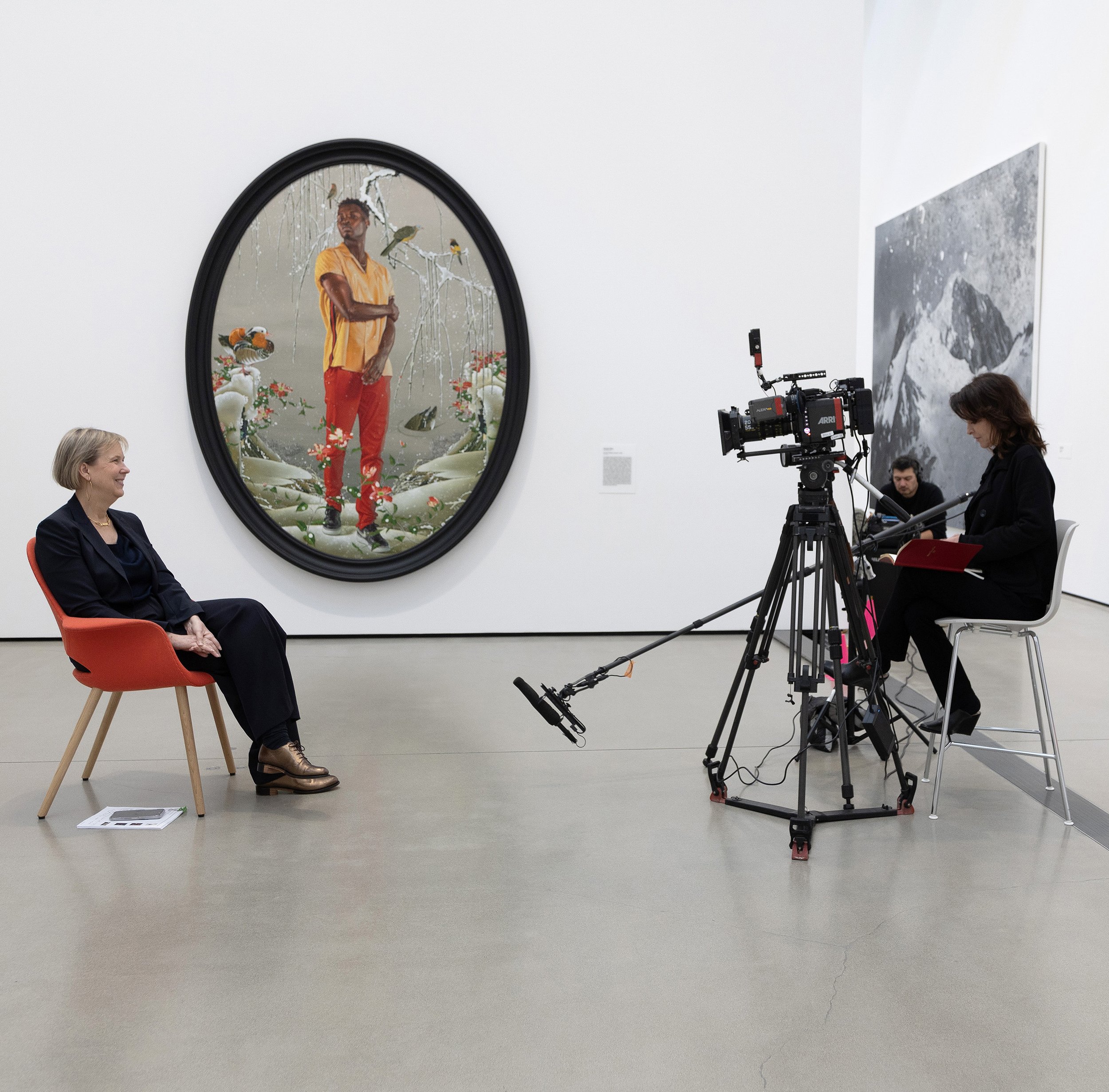 a couple people behind a camera interviewing a person in a chair with two paintings on the wall