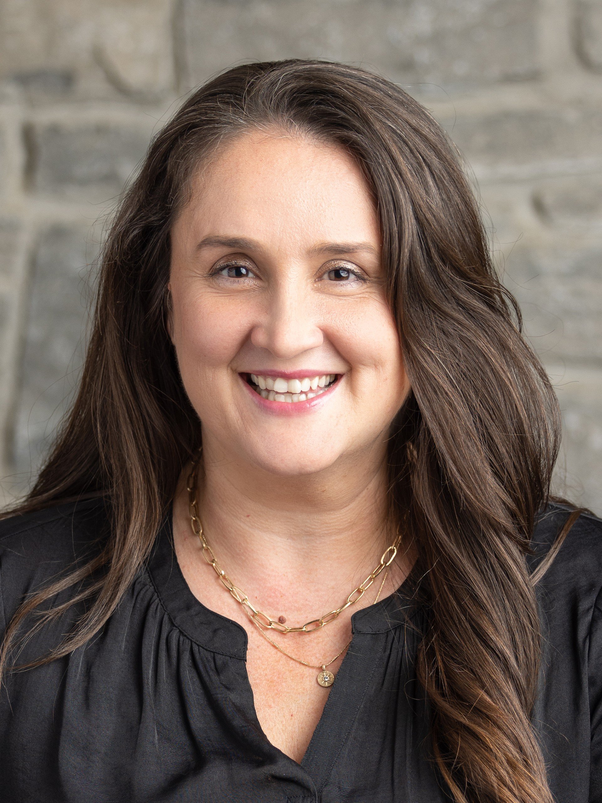 Close-up of a smiling woman with long brown hair wearing a black top and layered gold necklaces, standing in front of a gray brick wall.
