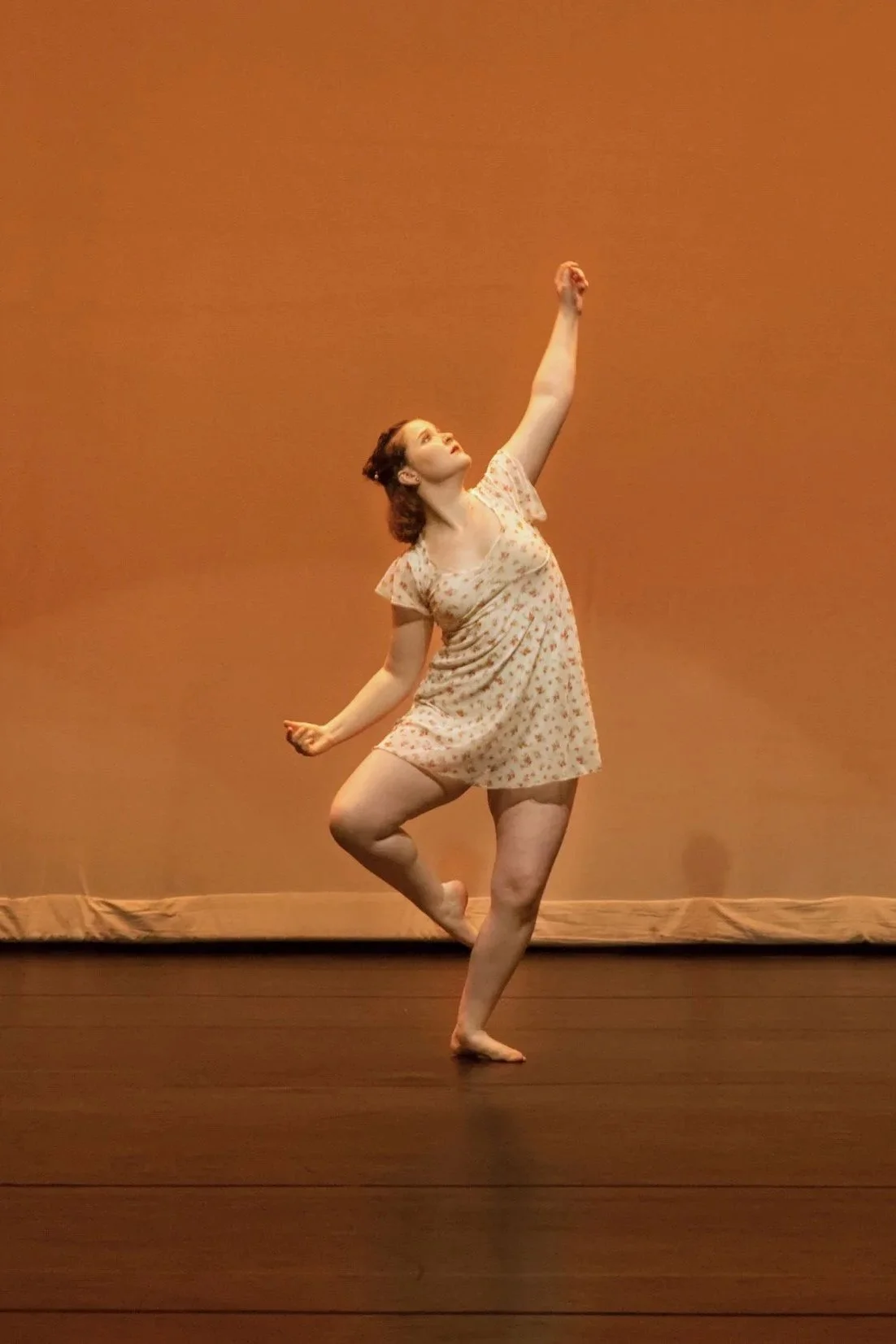 A woman dancing on stage, wearing a short, floral nightgown, against a plain background.