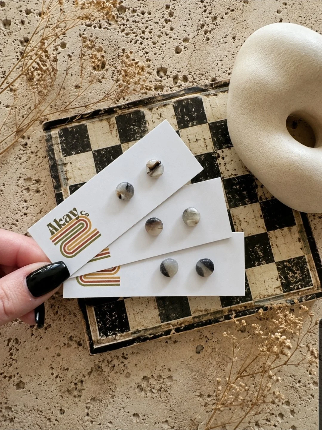 Small round polymer clay stud earrings in grey and black marble, displayed on Akay Collective branded cards with a vintage checkerboard box and dried botanicals
