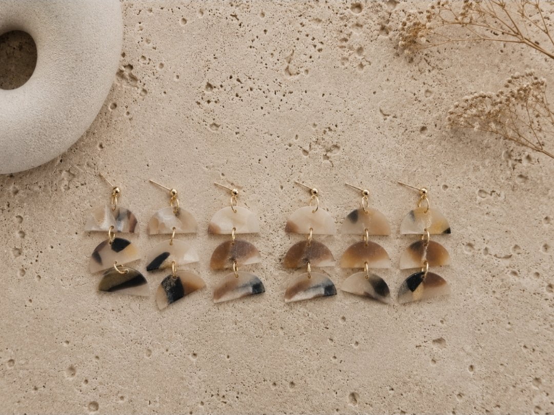 Two-inch cascading triple half-moon drop earrings in tortoise polymer clay with gold ring connectors and gold-filled ball posts, displayed on a travertine surface with dried botanicals