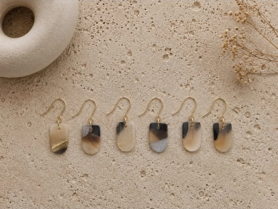 One-inch tortoise arch drop earrings in cream, black, and grey polymer clay with gold-filled ear hooks, displayed in a row on a travertine surface