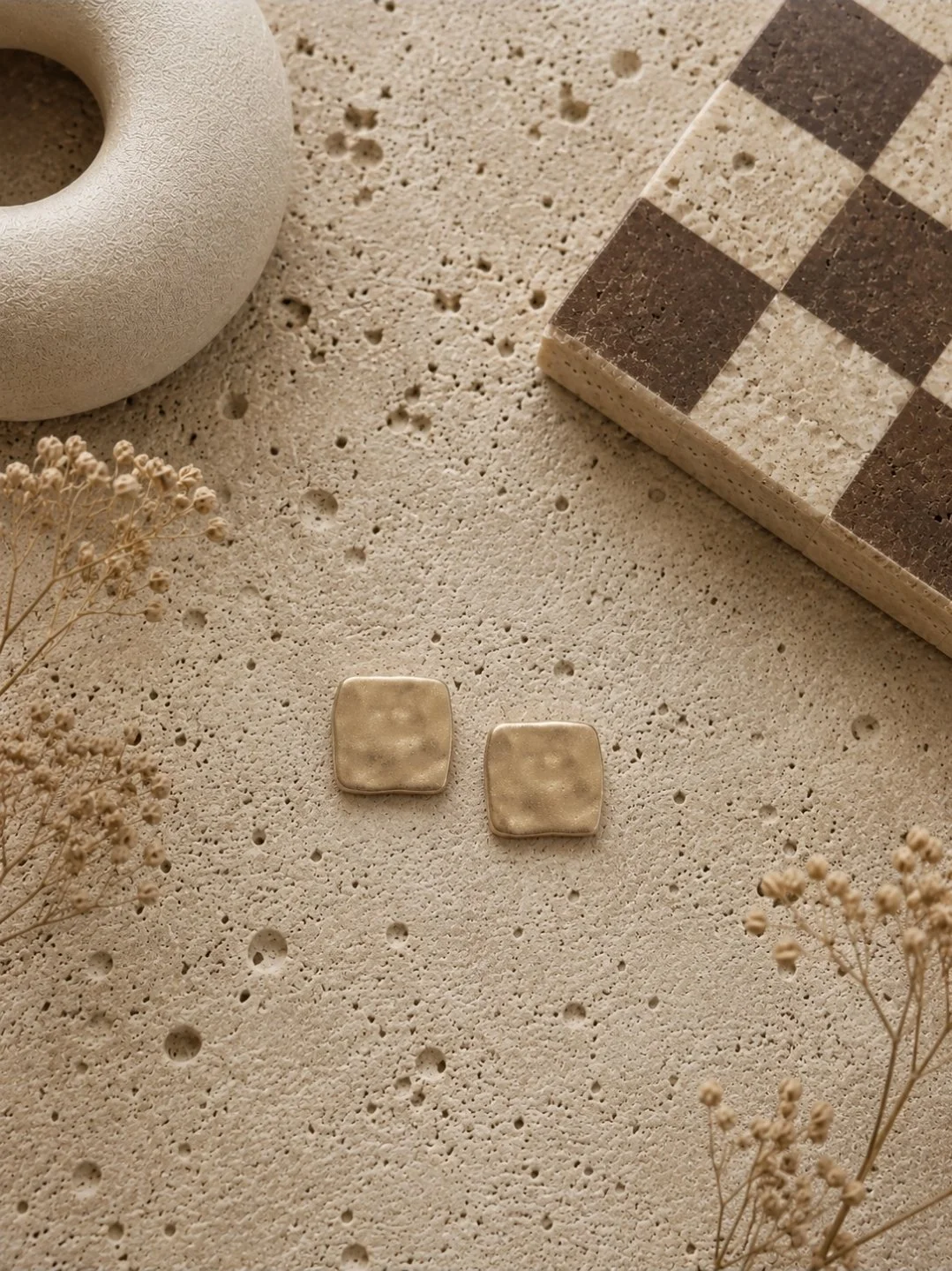 Half-inch hammered gold square stud earrings in glossy antique gold polymer clay, displayed on a travertine surface with dried botanicals and a checkerboard stone box