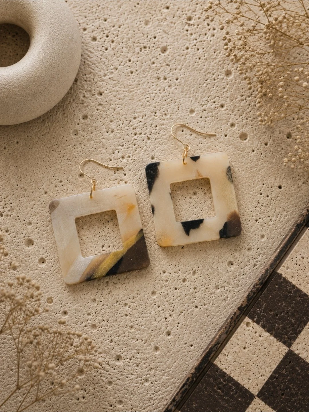 One and a half inch open square drop earrings in tortoise polymer clay with a cutout center, cream, black, and gold swirl, with gold-filled ear hooks on a travertine surface