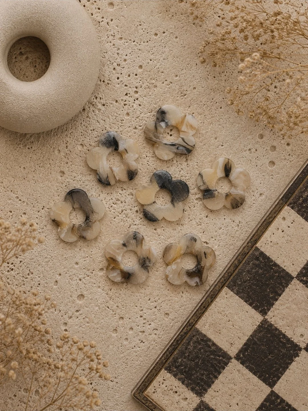 One-inch glossy tortoise flower stud earrings in black, cream, and gold polymer clay, displayed on Akay Collective branded cards with a checkerboard tray and neutral textures