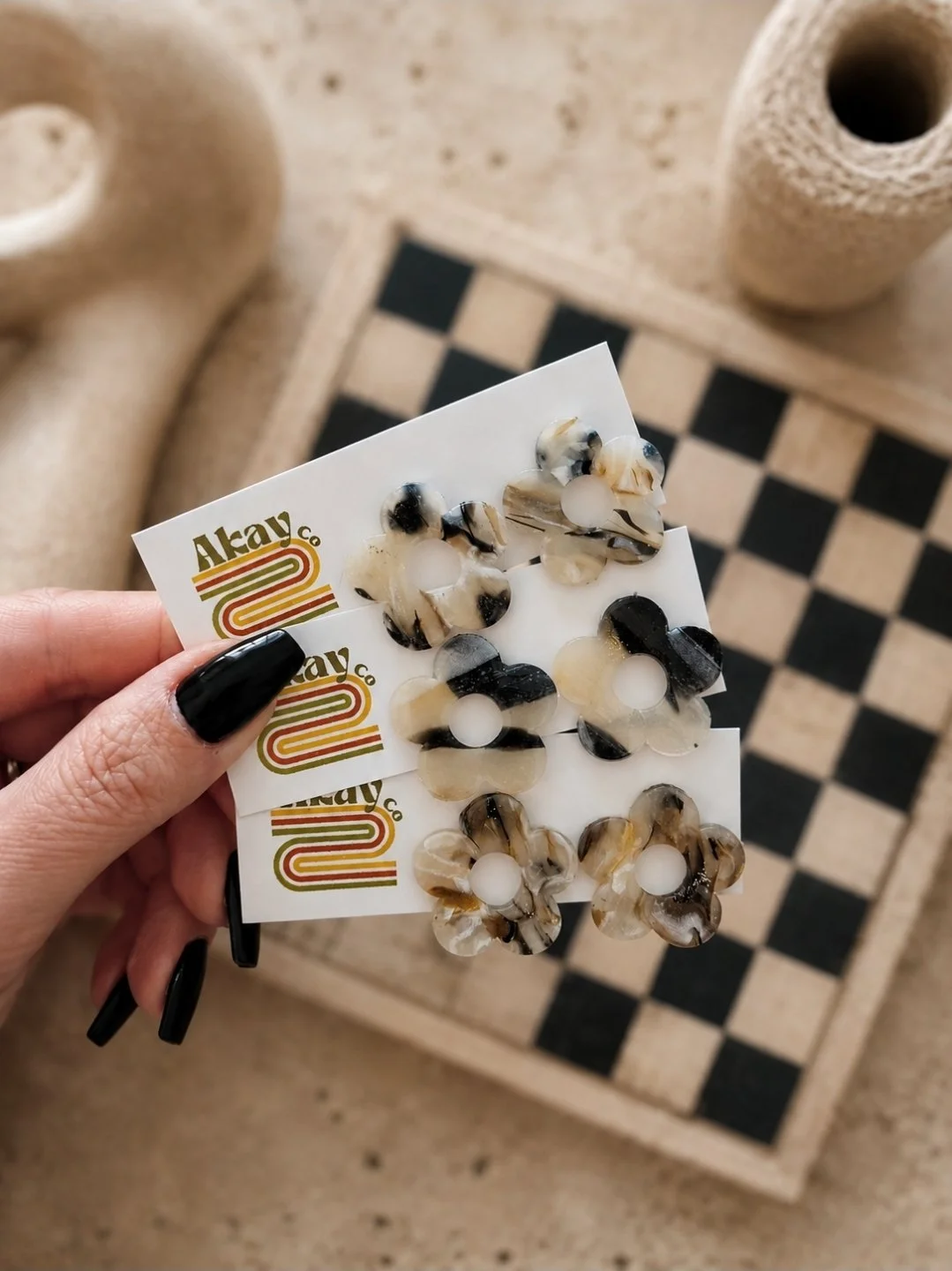 One-inch glossy tortoise flower stud earrings in black, cream, and gold polymer clay, displayed on Akay Collective branded cards with a checkerboard tray and neutral textures