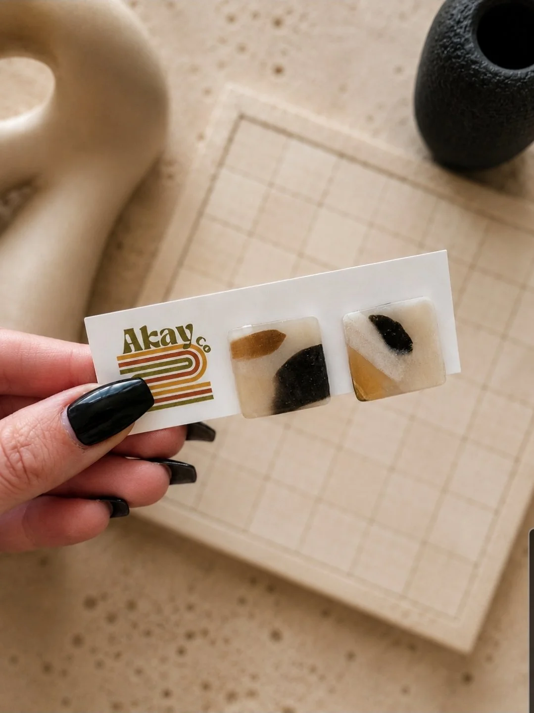 Three-quarter inch glossy tortoise square stud earrings in black, cream, and gold polymer clay, displayed on an Akay Collective branded card with neutral textured props
