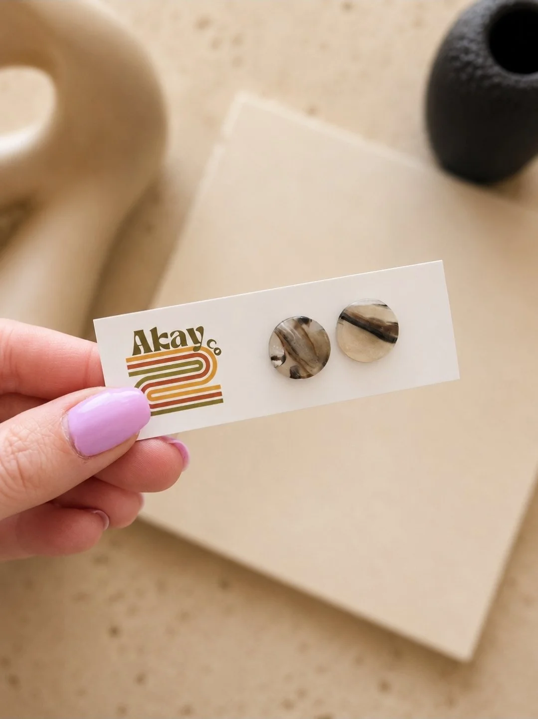 Half-inch glossy tortoise circle stud earrings in warm black, cream, and brown polymer clay, displayed on an Akay Collective branded card with neutral ceramic and linen props