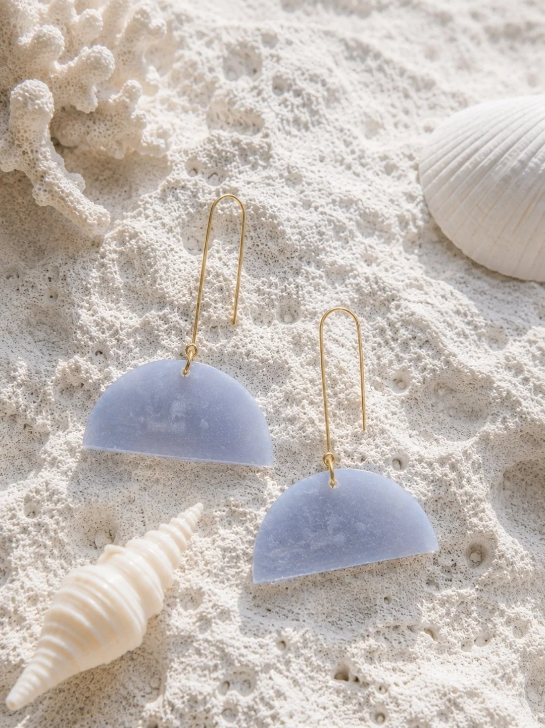 Two-inch frosted periwinkle blue half-moon polymer clay dangle earrings on gold-filled threader hooks, displayed on white sand with seashells and coral