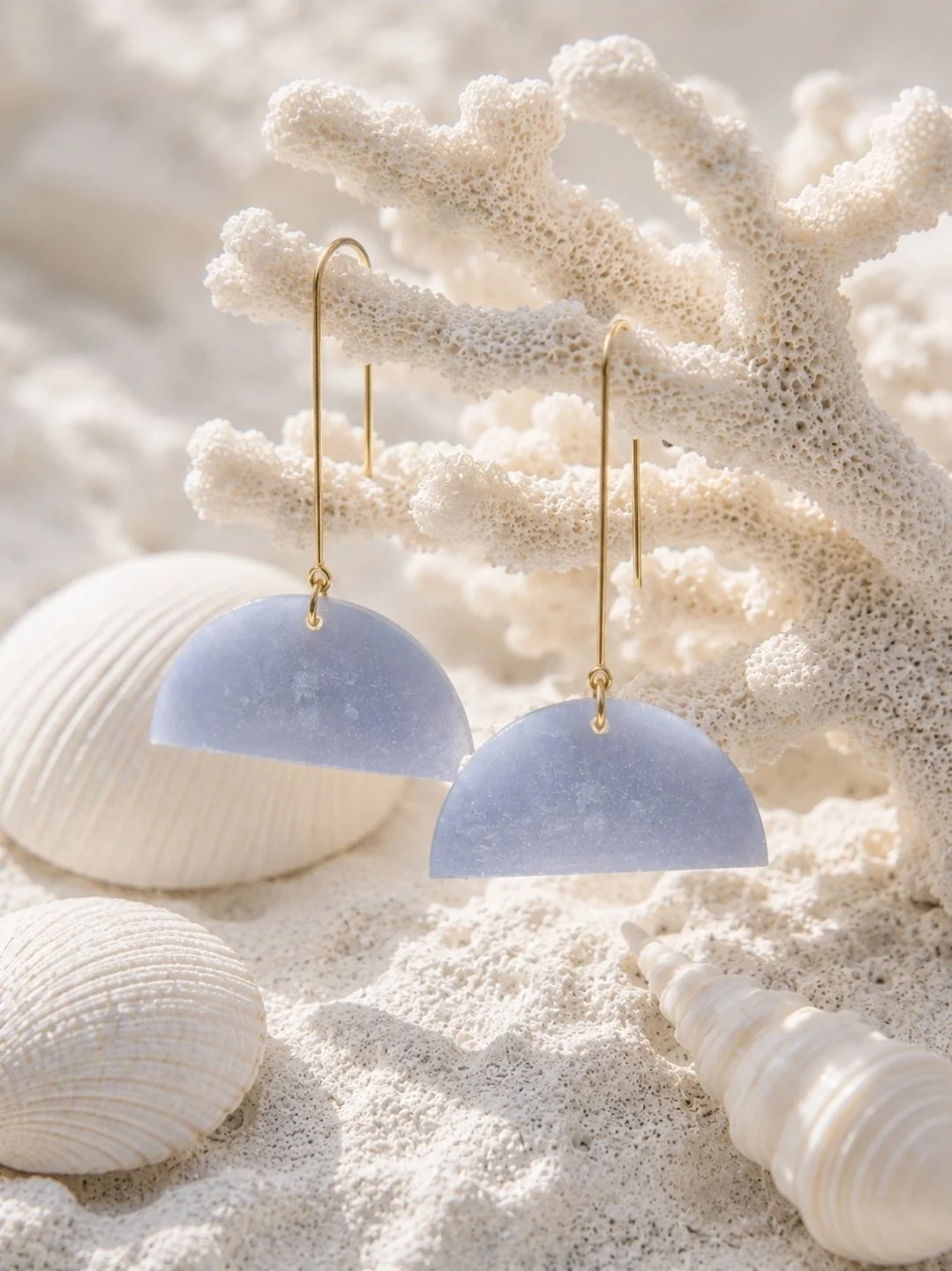 Two-inch frosted periwinkle blue half-moon polymer clay dangle earrings on gold-filled threader hooks, displayed on white sand with seashells and coral