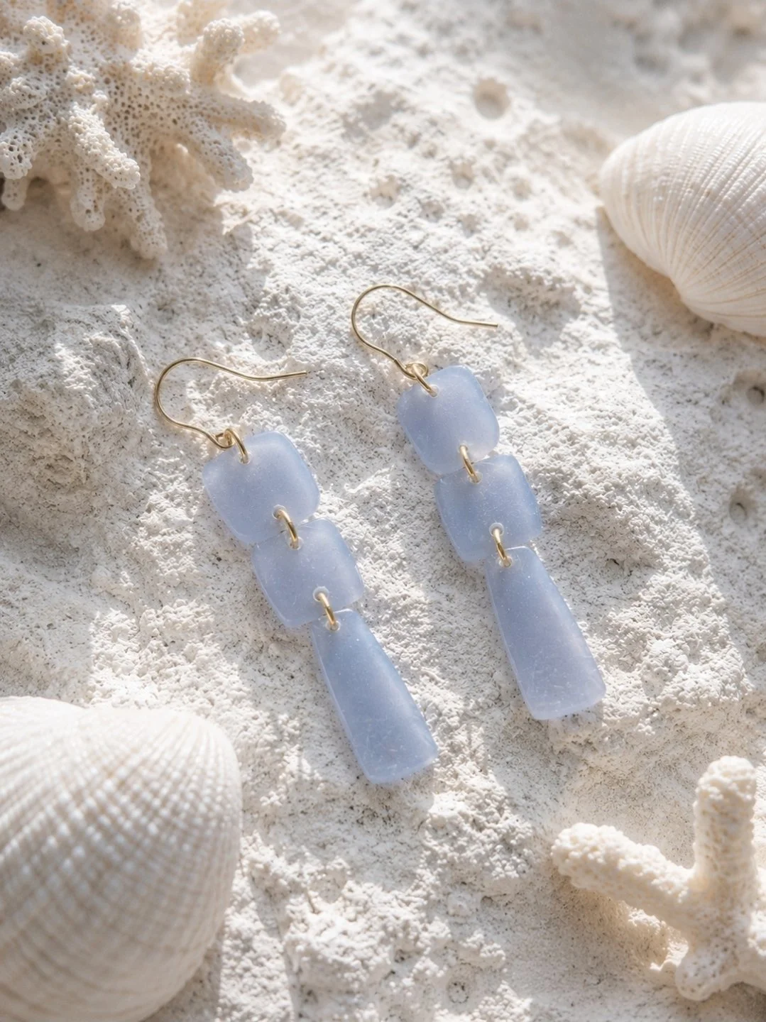 Two and three-quarter inch frosted periwinkle blue polymer clay drop earrings with three linked pieces and gold-filled hooks, displayed on white sand with seashells and coral