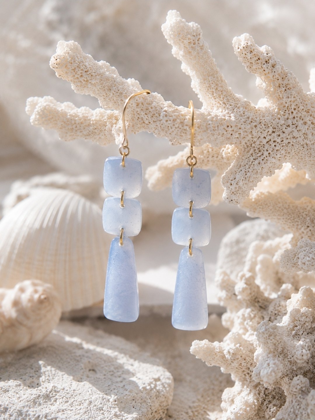 Two and three-quarter inch frosted periwinkle blue polymer clay drop earrings with three linked pieces and gold-filled hooks, displayed on white sand with seashells and coral
