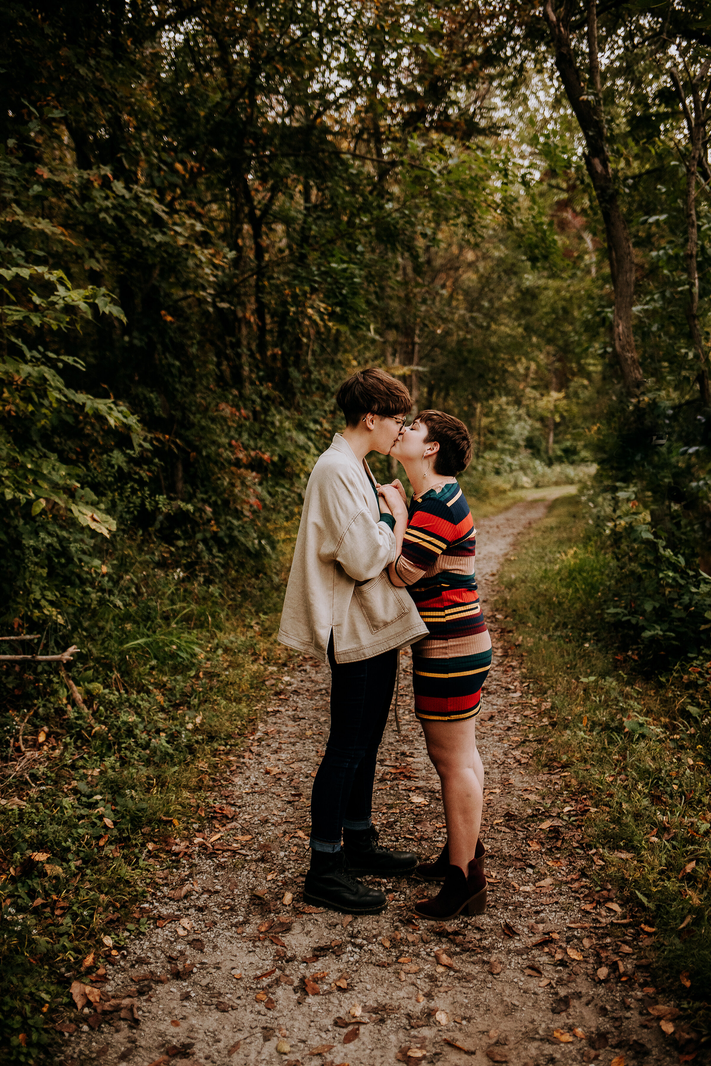  Beautiful same-sex couple kissing on a tree-lined trail wearing a striped dress and boots and a tan coat by Kindred + Co in Indiana. fall engagments outdoor photography indiana locations for outdoor engagments same-sex couple pose inspo fall outfits