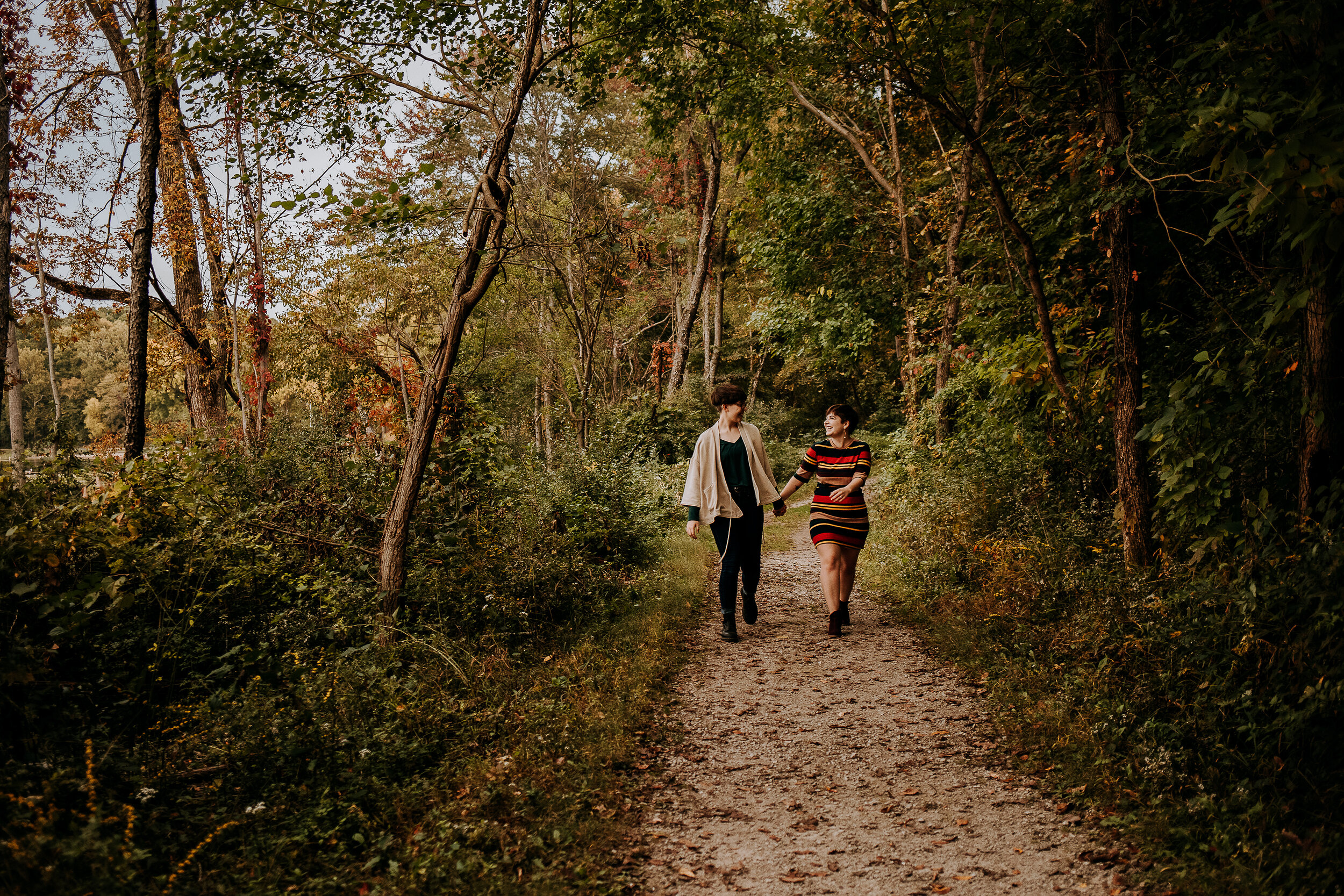  Candid shot of same-sex couple holding hands and walking on an outdoor trail with trees in a fall striped dress by Kindred + Co in Indiana. boho wedding photographer same-sex couple engagement photos pose inspo fall engagement photo outfit inspo dre