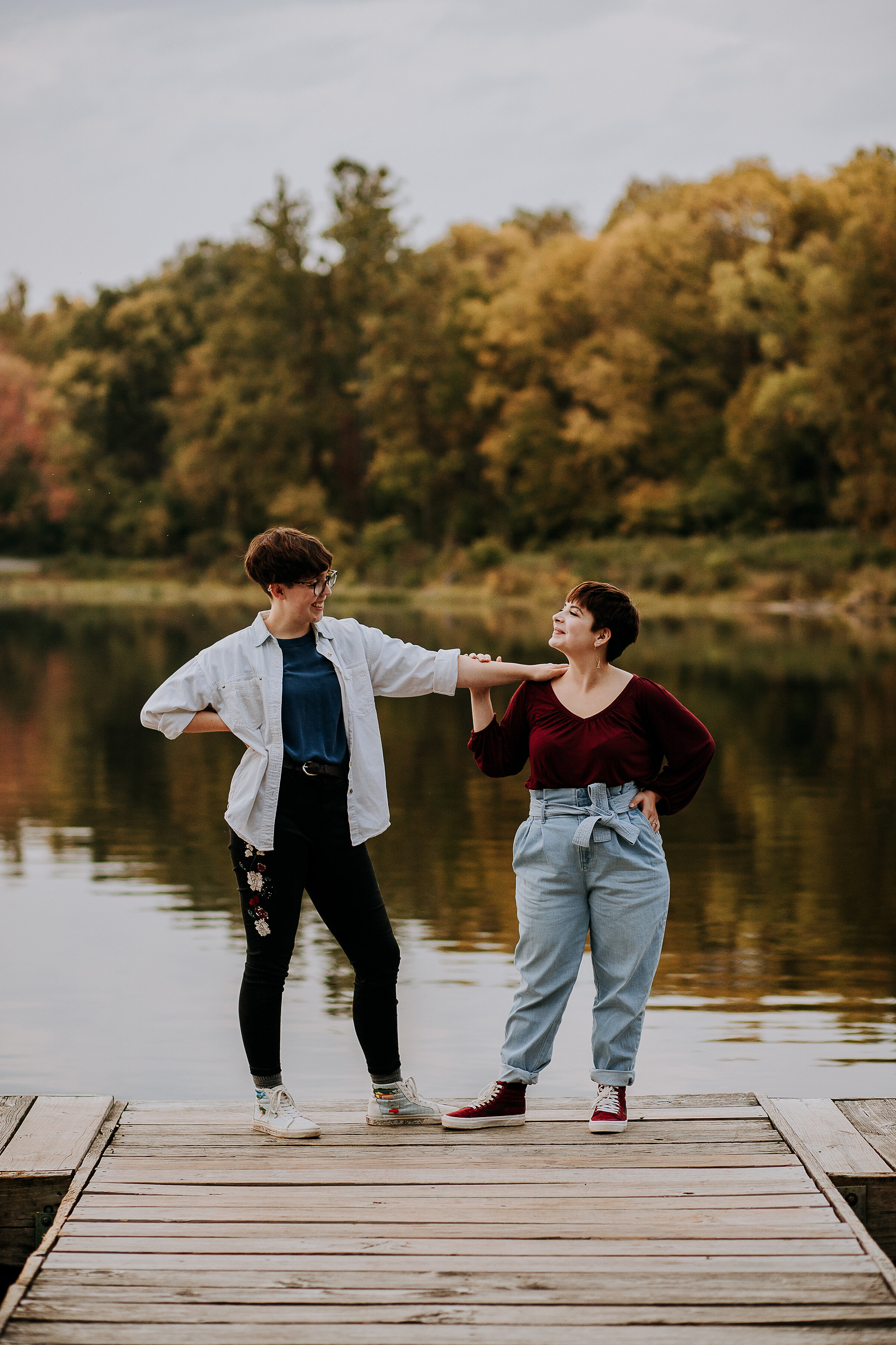  Candid photo of same-sex couple on a dock by a lake in the fall wearing maroon and gray in Indiana by Kindred + Co. lake and dock location for photoshoot in indiana fort wayne indiana same-sex couple pose inspo for engagements casual fall outfits wi