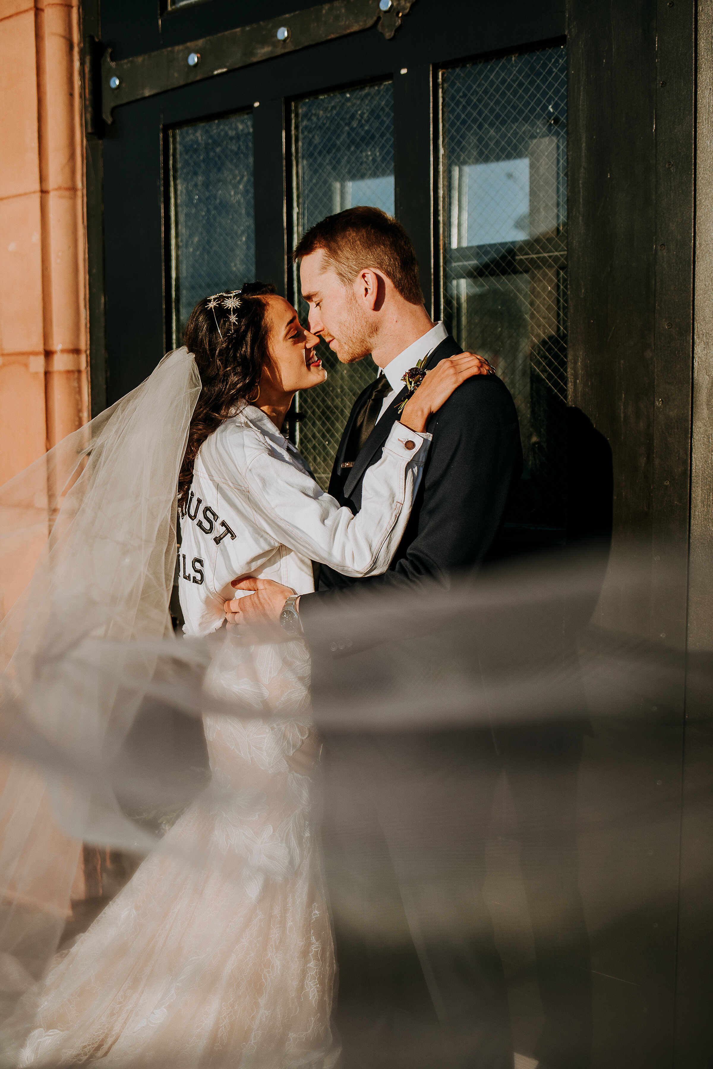  With Kindred &amp; Co. Photography capturing their wedding portraits, this newlywed bride and groom lean in for a kiss at Eastern Market Michigan. white denim jacket on bride, professional wedding photographer downtown detroit michigan, hip length w