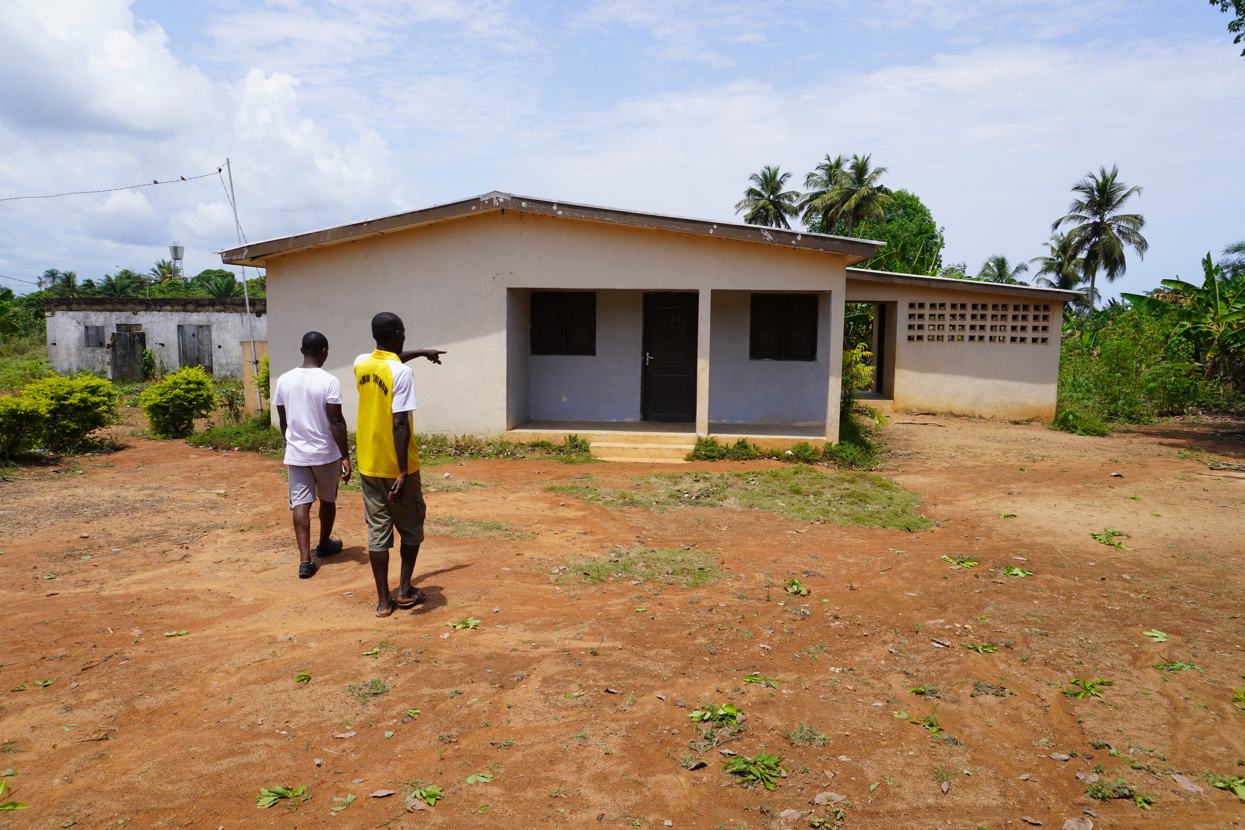  Alex Nabo montre la maison de la directrice de l’école, en déplacement, construite aussi dans le cadre du “Projet village”. Au fond, à gauche, on peut voir le château d’eau financé par la coopération espagnole.  