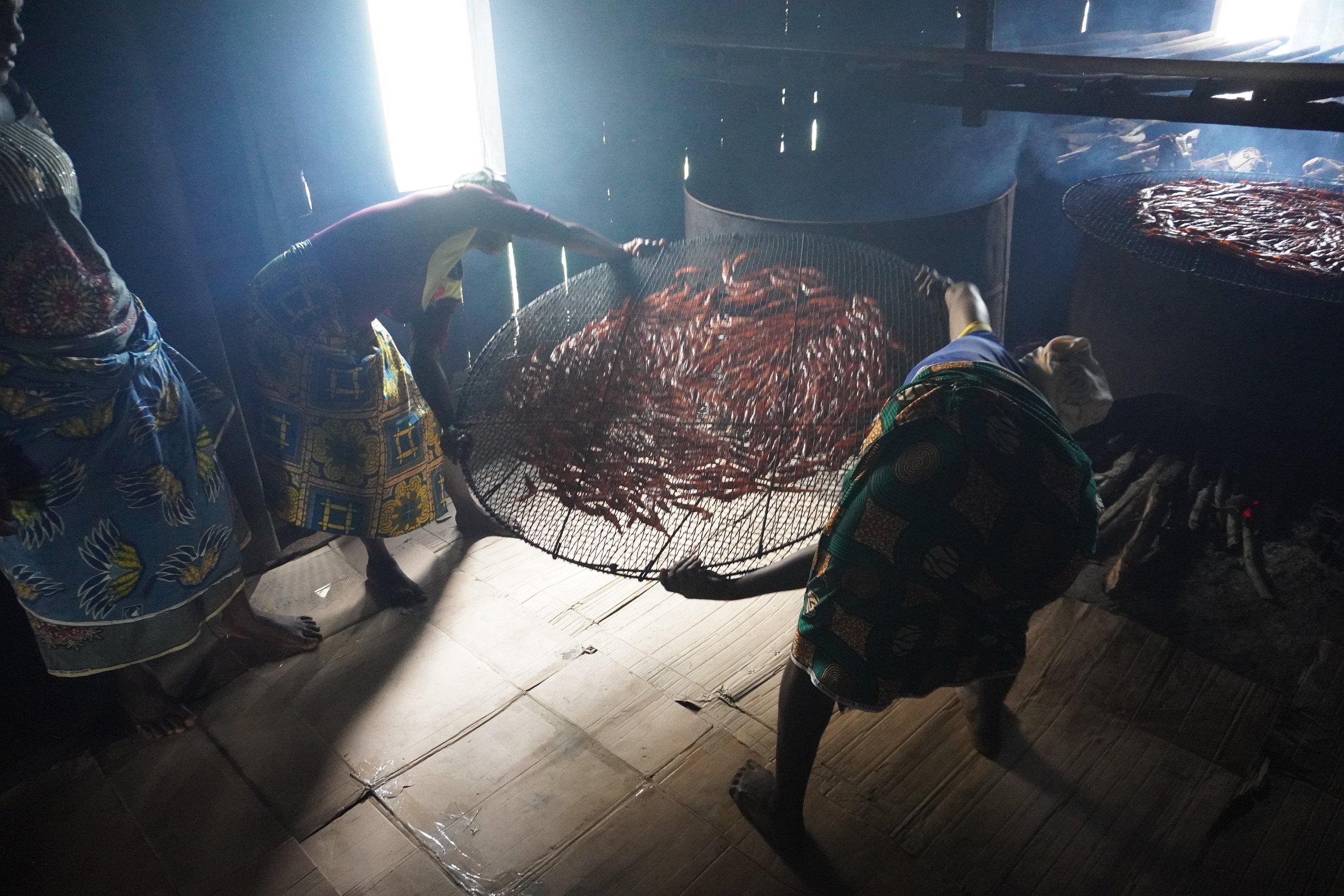  Eugénie Zannou et Pierrette Ahouandjinou, deux femmes de la coopérative de mareyeuses et transformatrices de crevettes (COMATRANC) sur le Lac Nokoué retournent les crevettes qu’elles fument sur des fours traditionnels. La maison sur pilotis, en bois