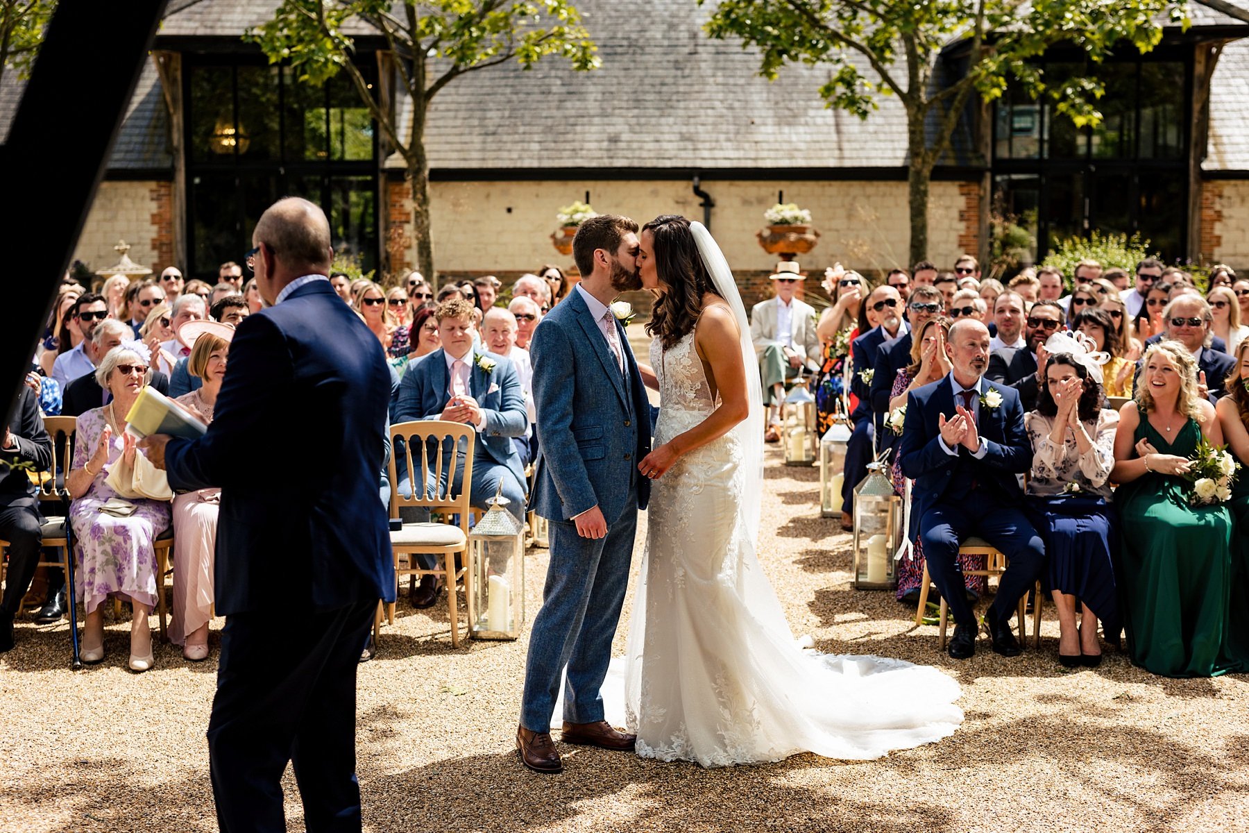 A bride and groom kiss as their guests look on in the background at their wedding ceremony