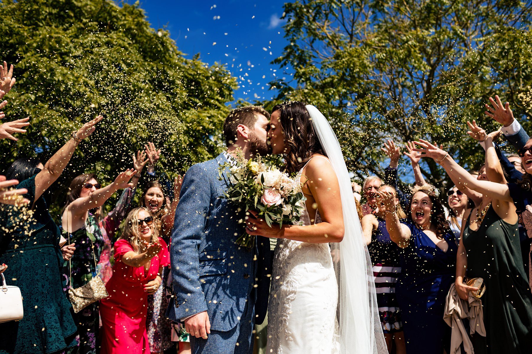 A bride and groom kiss as they are showered with confetti by their surrounding guests at their wedding at Bury Court Barn