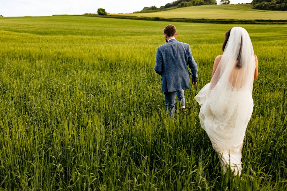 A bride and groom walk through tall grass after their wedding at Bury Court Barn