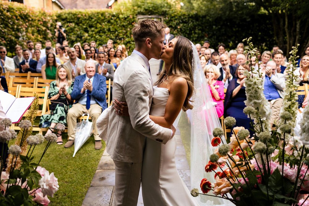 A bride and groom kiss at their outdoor wedding at Millbridge Court, as the guests in the background look on and cheer.