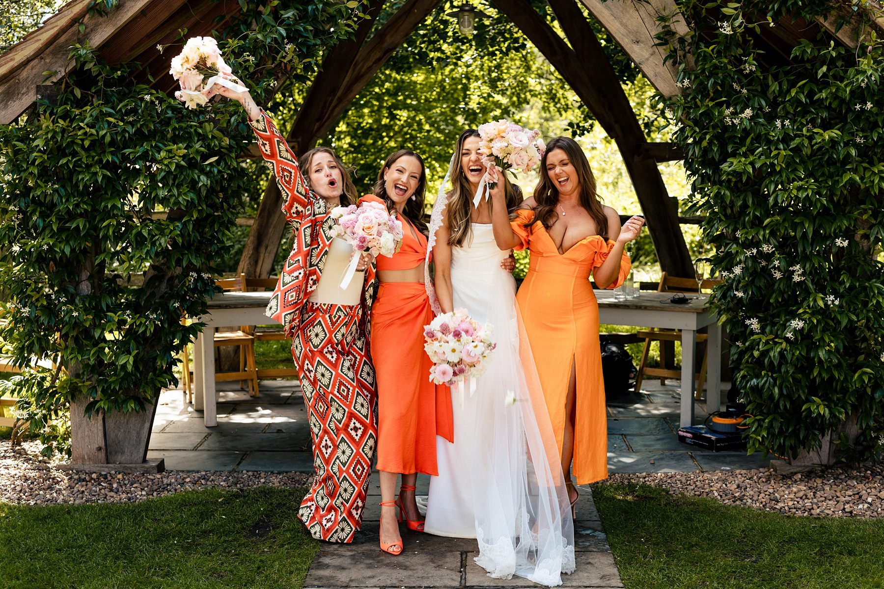 A bride and her bridesmaids cheer and laugh at Millbridge Court, the bride wearing white and the bridesmaids in warm orange outfits.